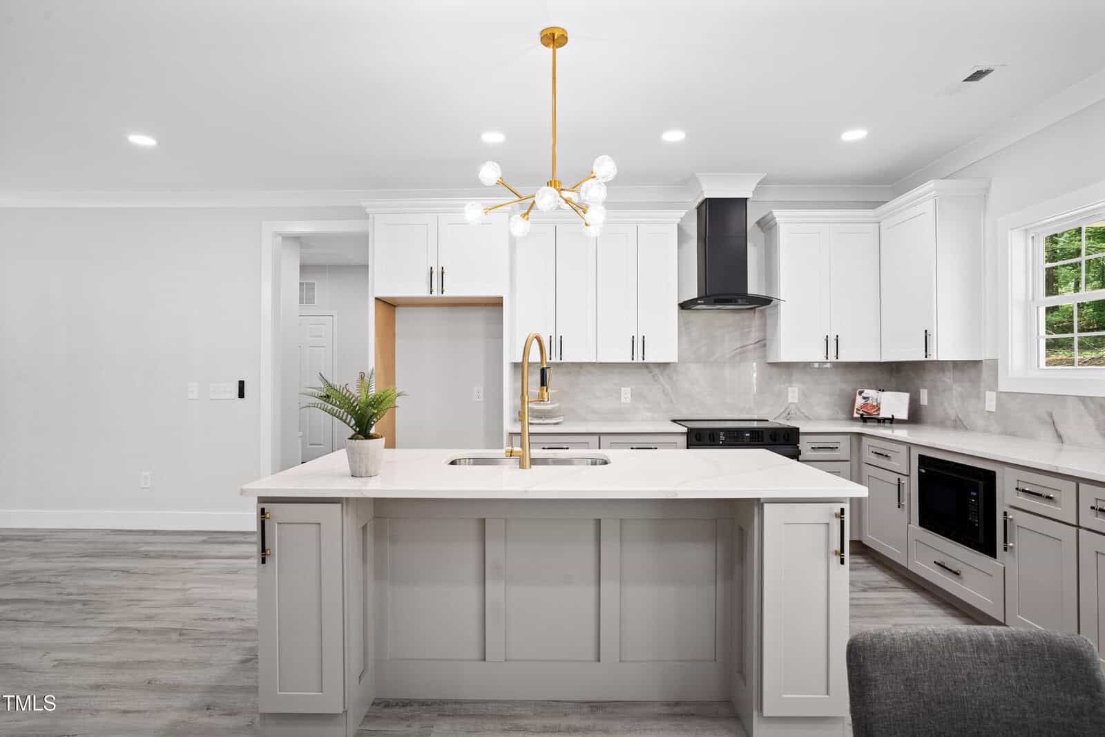 Modern kitchen with white cabinets, gray island with sink, gold faucet, stainless steel appliances, marble backsplash, and a contemporary chandelier. Large window on the right lets in natural light. Light wood floors throughout.