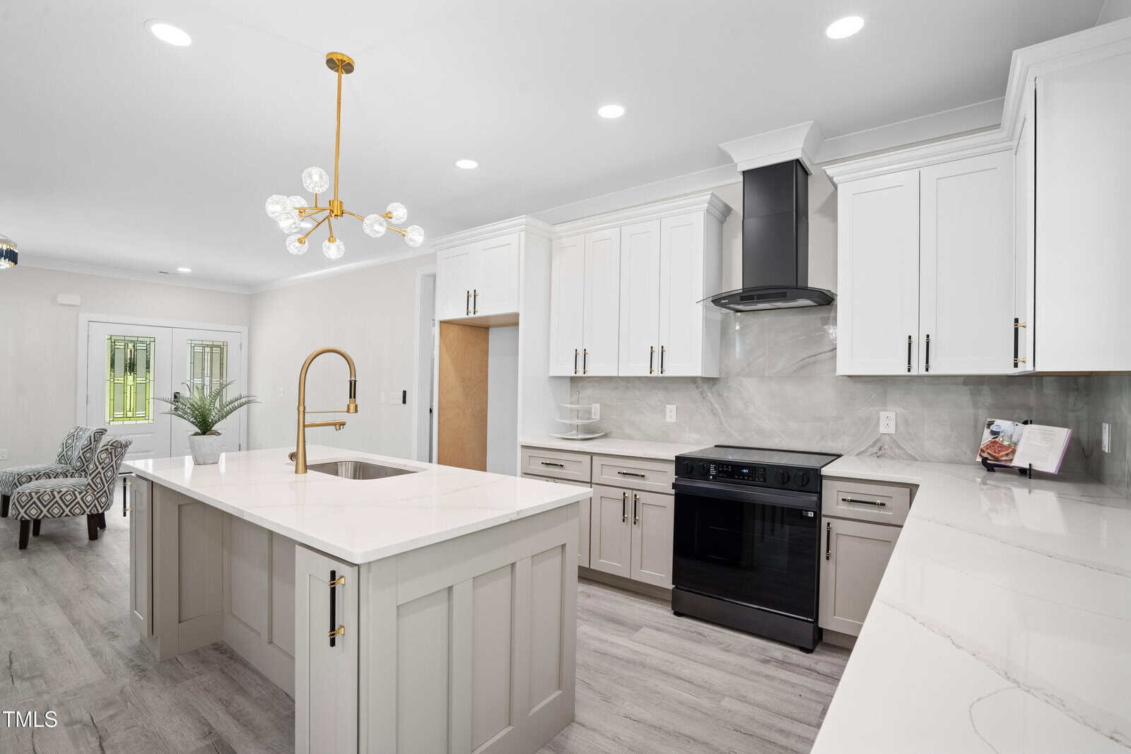 Modern kitchen with white cabinets, a large central island with a gold faucet, black appliances, light gray wood floors, and a stylish chandelier. An open doorway reveals a sitting area with two patterned chairs and large windows.