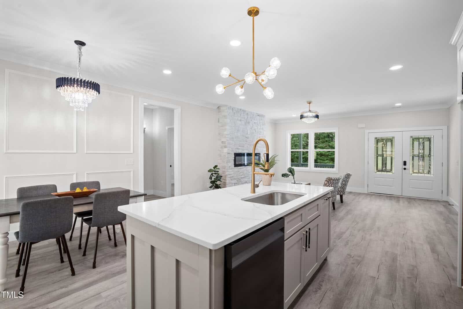 Modern open-concept kitchen and dining area with white walls, marble island, gray cabinets, gold light fixtures, and large windows letting in natural light. Dining table with six chairs is visible near a stone accent wall.
