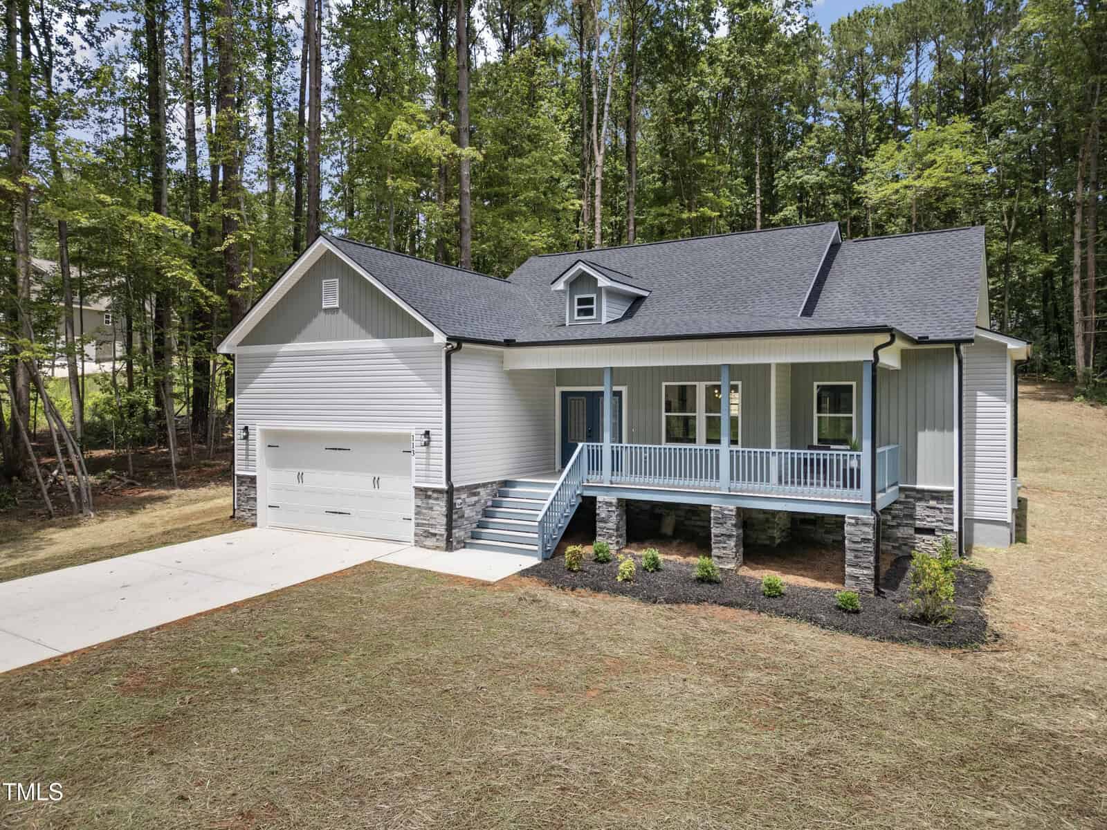 A modern single-story house with gray siding, a front porch with blue railings, stone accents, and a two-car garage, surrounded by grass and tall trees.