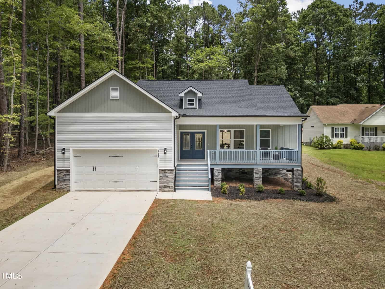 A modern single-story house with a gray and white exterior, double garage, front porch with railings, landscaped yard, and surrounded by trees and grass.