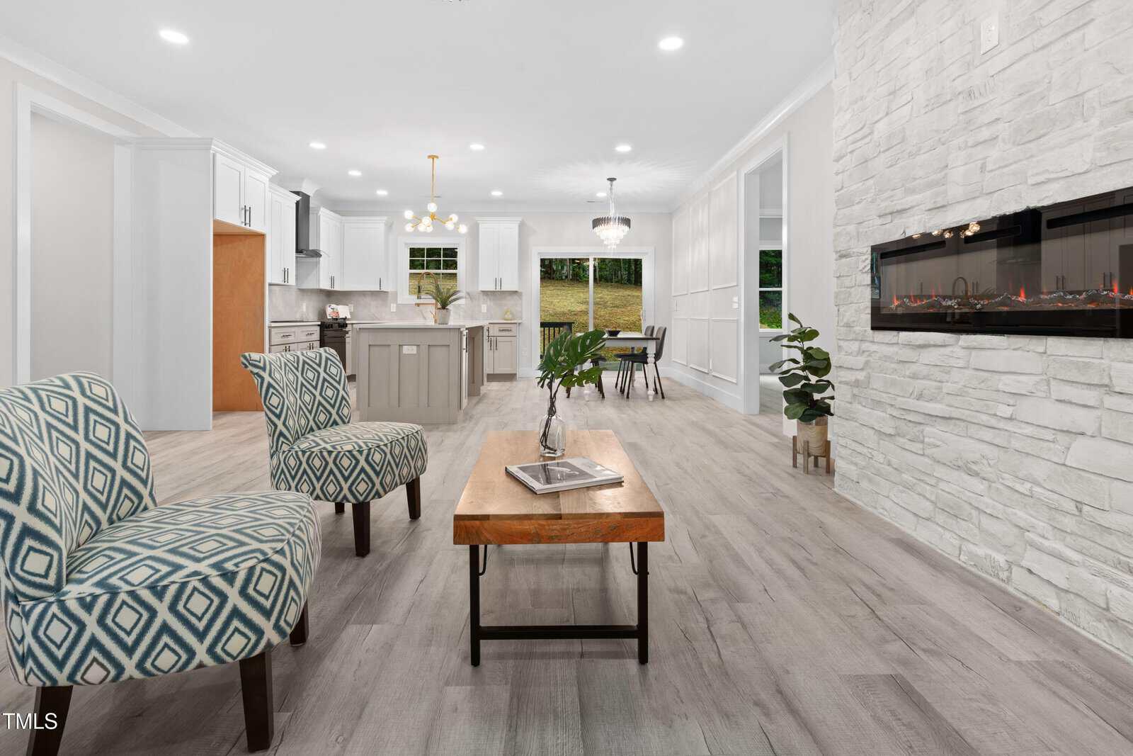 Modern open-concept living room with patterned chairs, a wooden coffee table, white stone fireplace, and view into a bright kitchen with white cabinets and an island. Large windows reveal greenery outside.