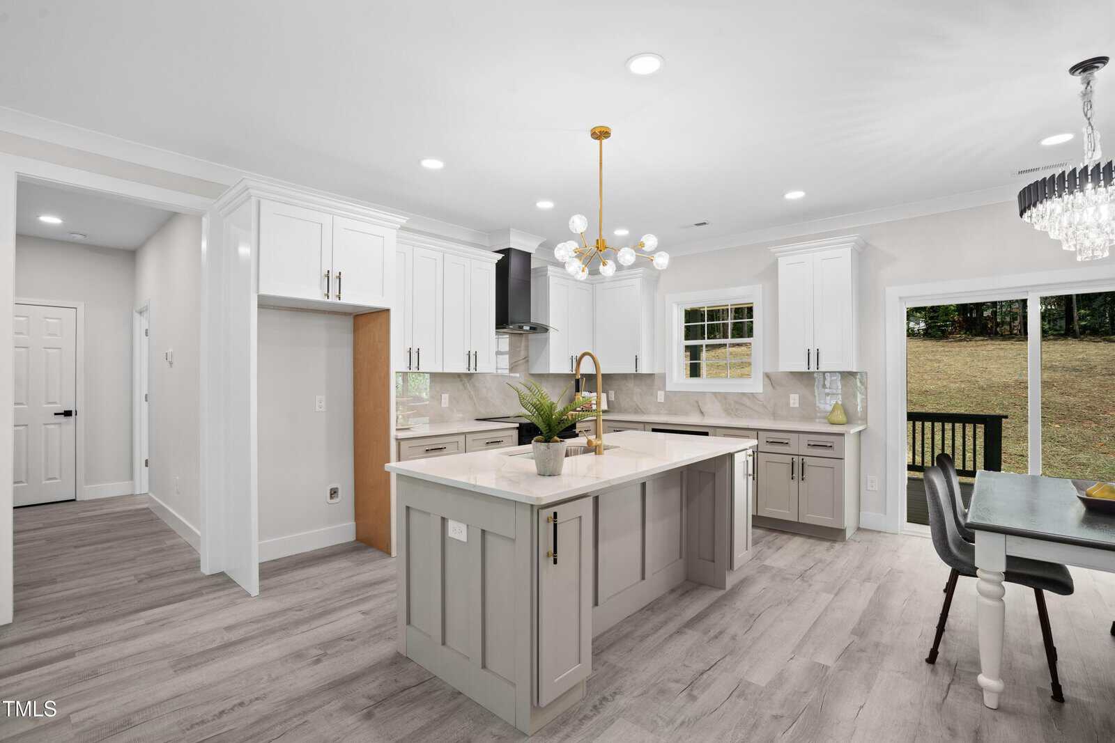 Modern kitchen with white cabinets, gray island, gold fixtures, light wood flooring, built-in appliances, and a chandelier. Dining area with table and chairs is adjacent to sliding doors leading to a backyard.