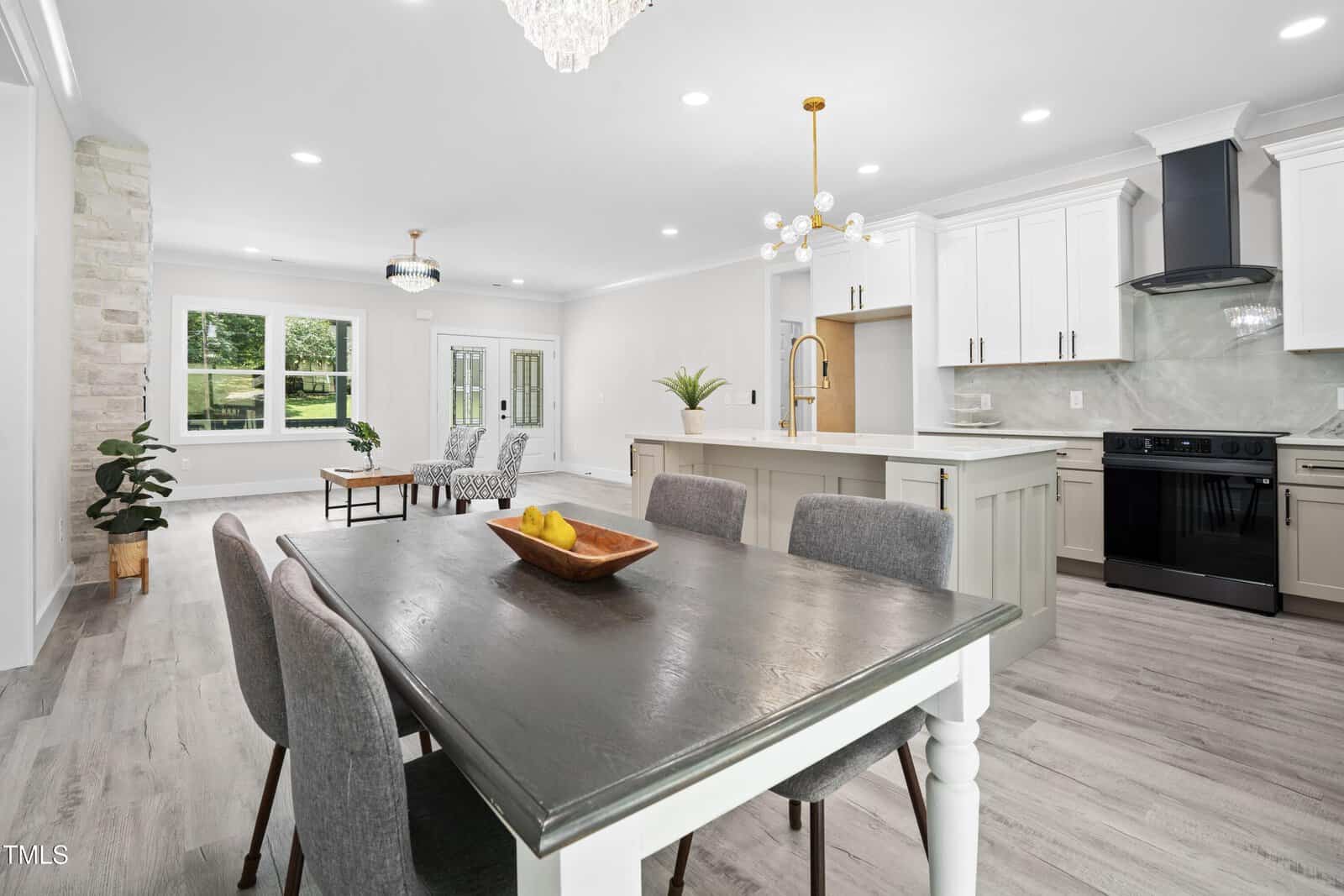 Modern open-concept kitchen and dining area with gray and white decor, marble backsplash, island with stools, chandelier, and large windows providing natural light. Living area with chairs is visible in the background.