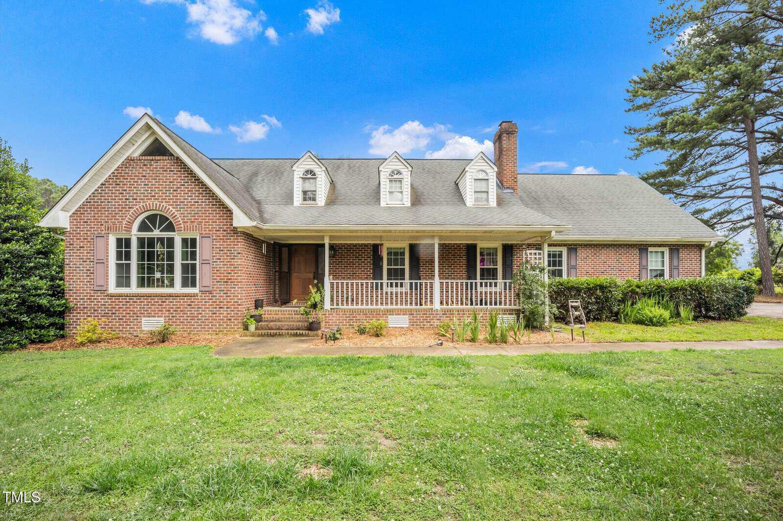 A single-story red brick house with a gabled roof, dormer windows, white shutters, and a covered front porch. The home has a large lawn, shrubs, and trees under a bright blue sky with scattered clouds.