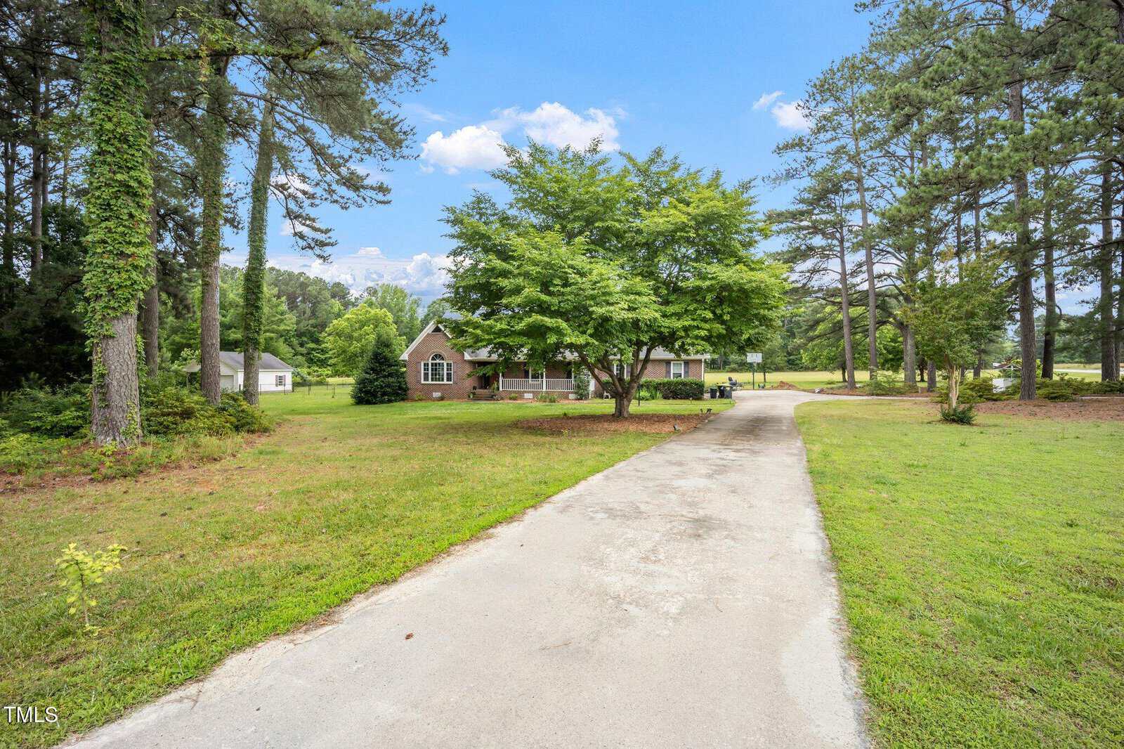A concrete driveway leads to a brick house with large windows, surrounded by tall trees and green lawn under a blue sky with clouds. A detached white garage is visible to the left.