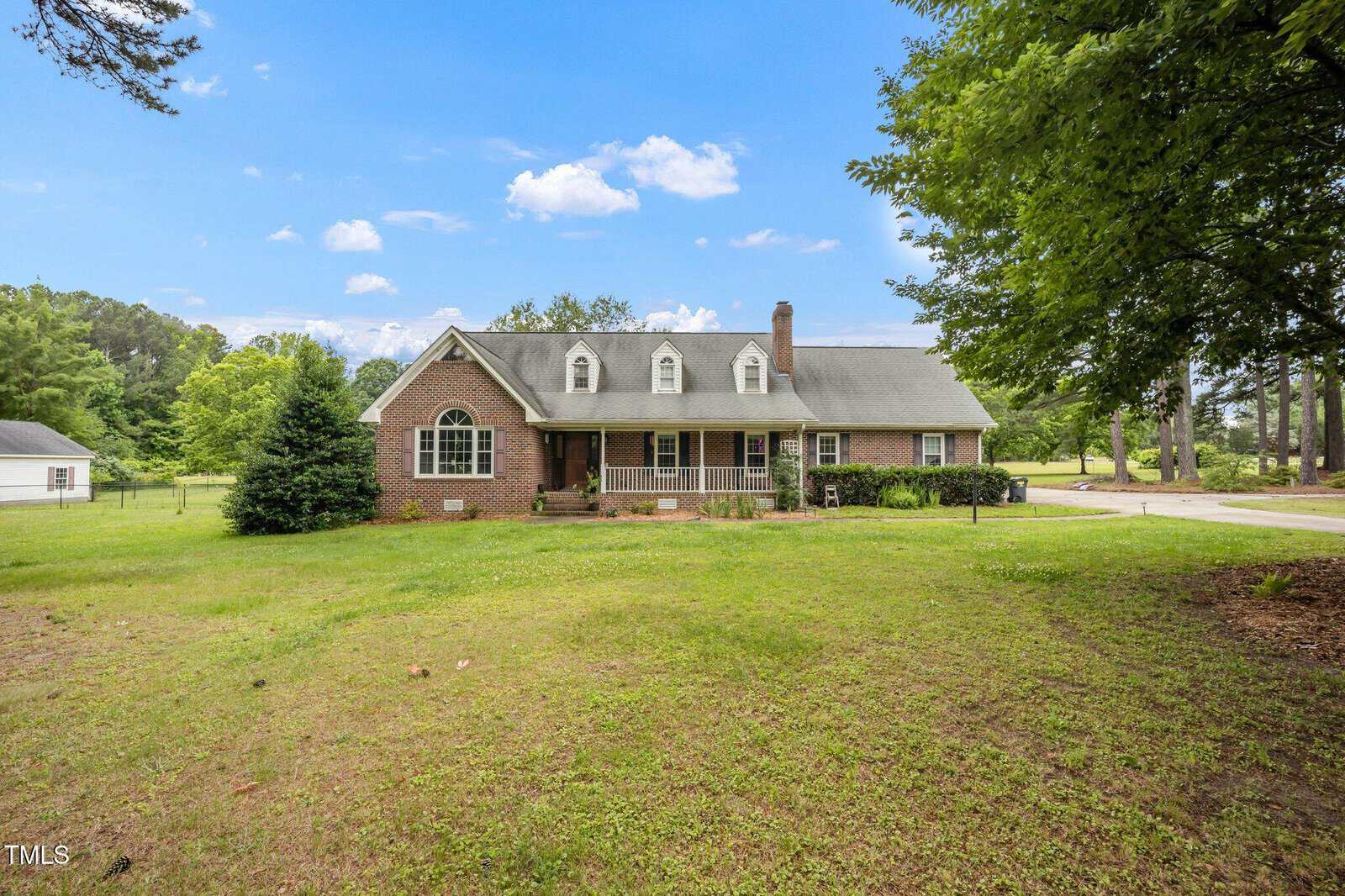 A brick house with dormer windows and a covered porch sits on a spacious, grassy lawn with large trees and a driveway, under a blue sky with scattered clouds.
