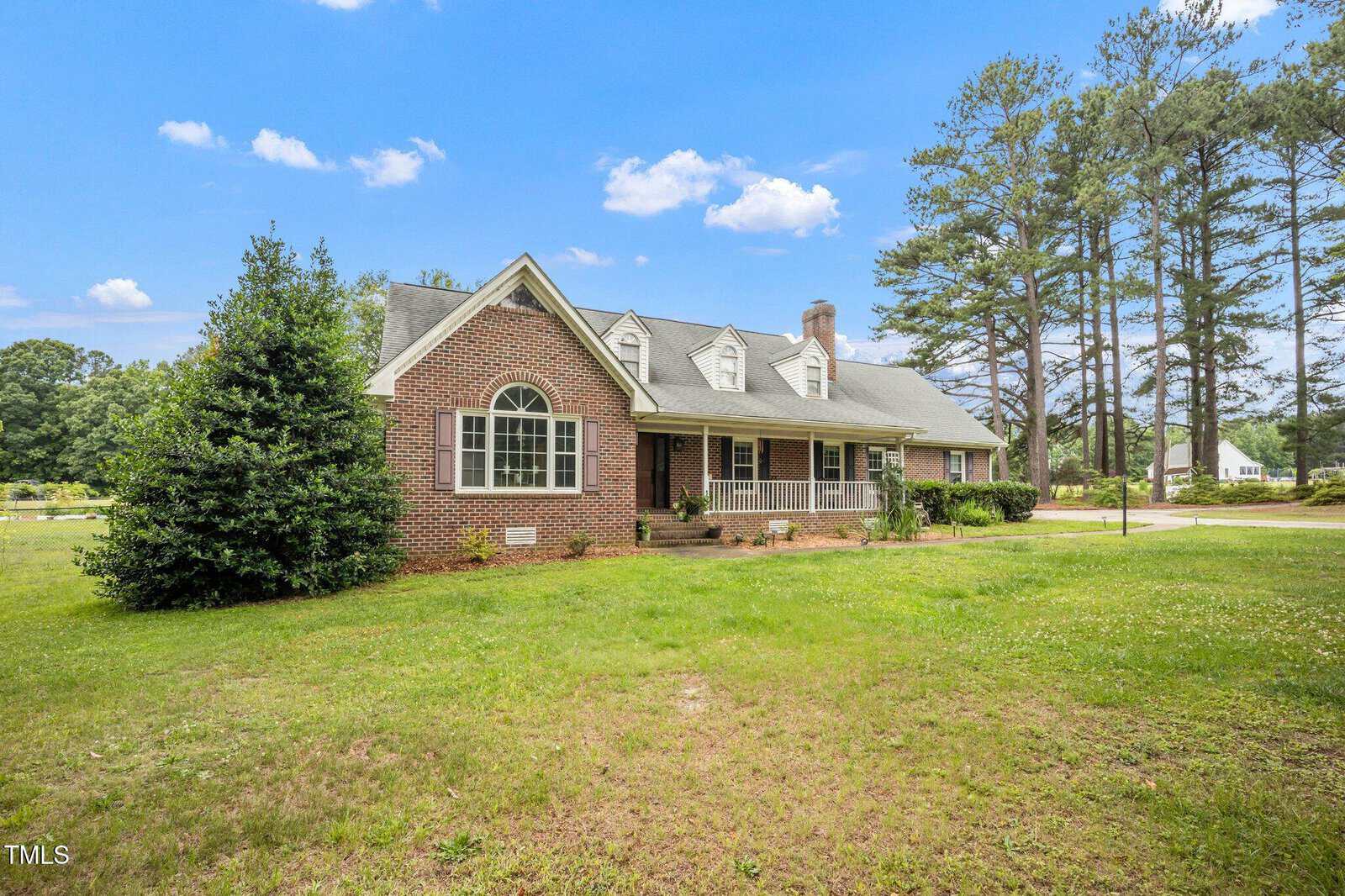 A brick house with gabled roof, arched window, and covered front porch sits on a large grassy lawn surrounded by tall pine trees under a blue sky with scattered clouds.