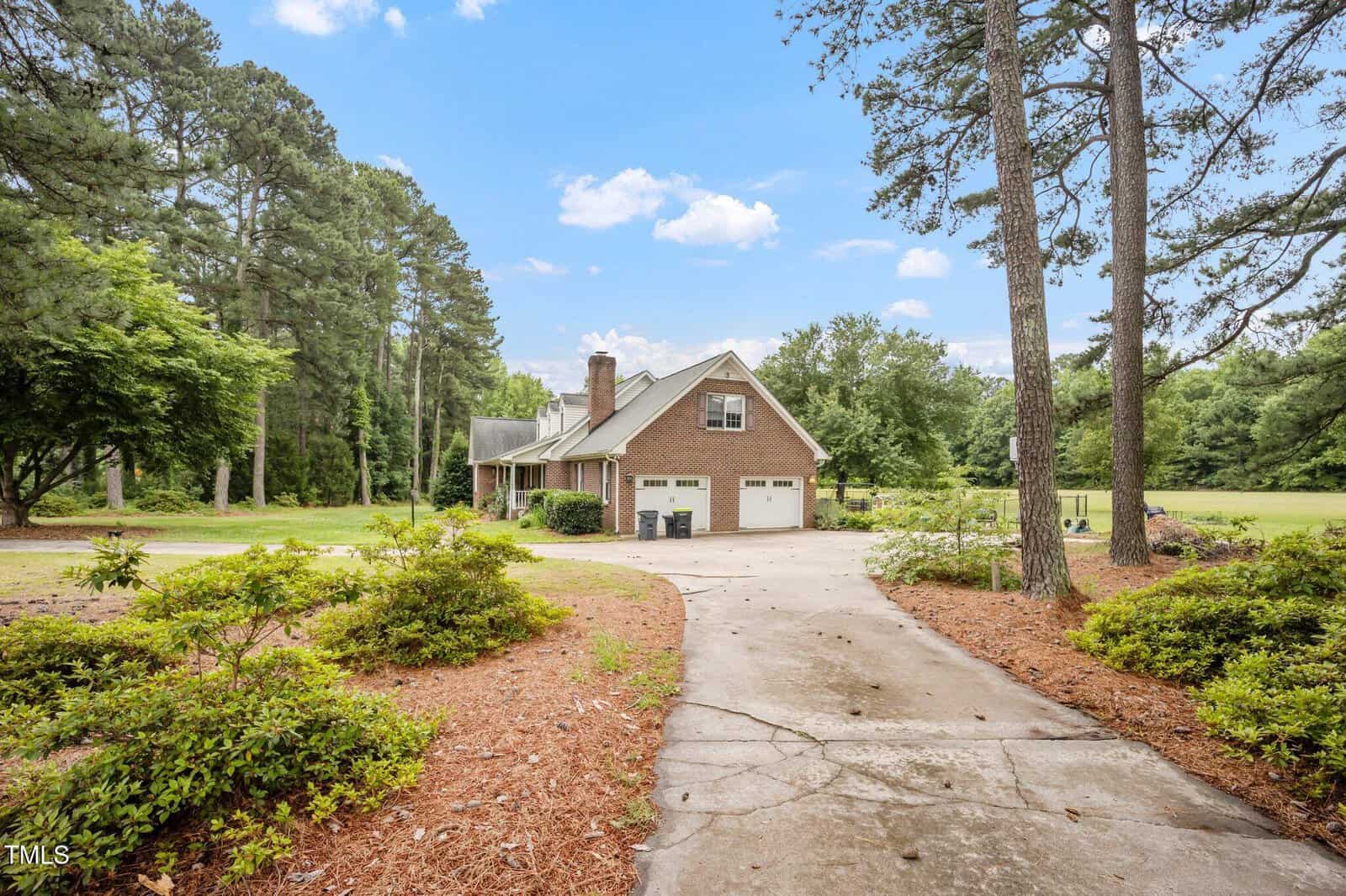 A brick house with a steep roof and two-car garage sits at the end of a curved driveway, surrounded by tall trees, shrubs, and a green lawn under a blue sky with scattered clouds.