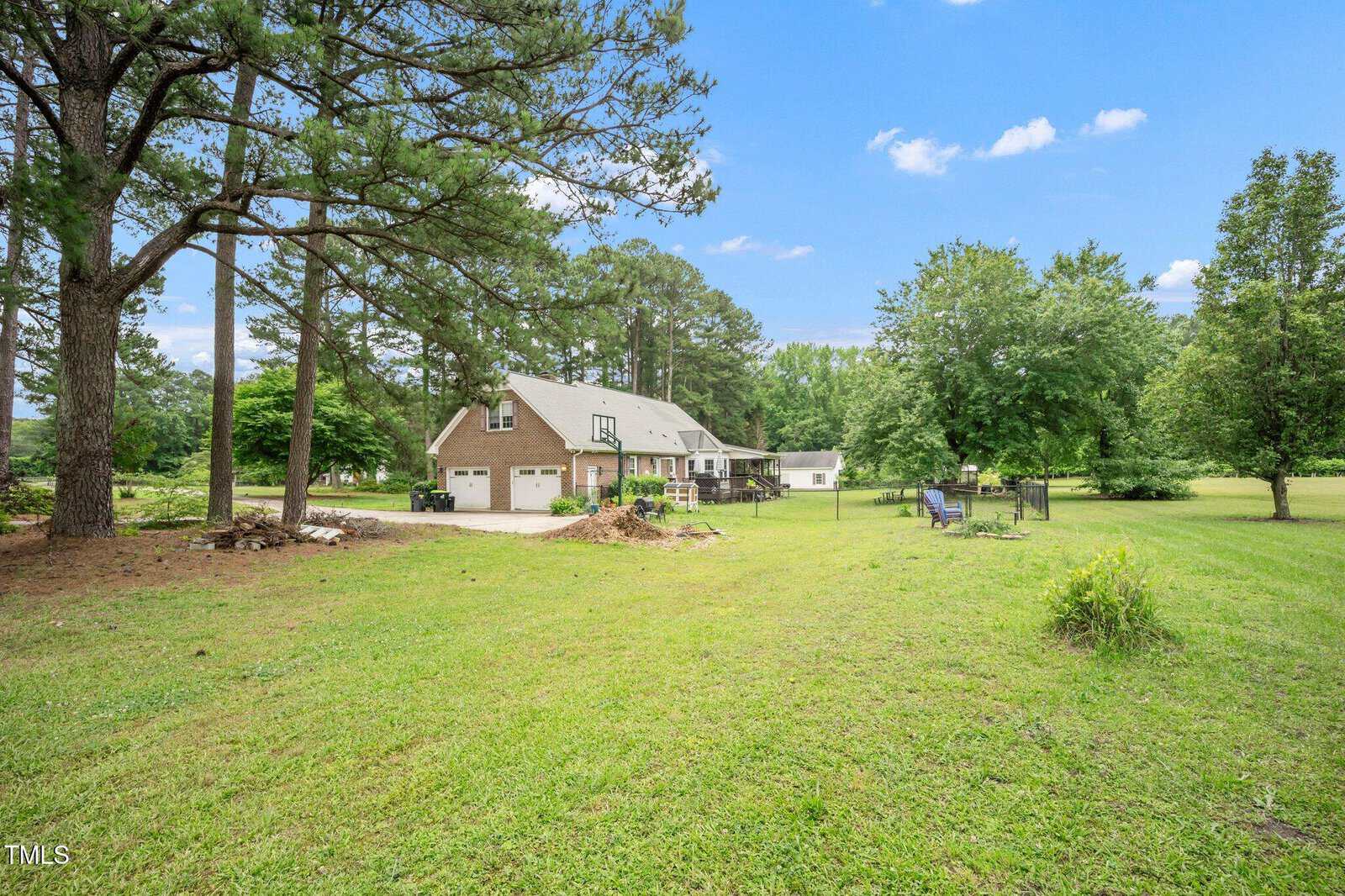 A large grassy yard with scattered trees, a two-story house with a garage in the background, lawn chairs with a firepit to the right, and a woodpile near the left edge. The sky is mostly clear with a few clouds.
