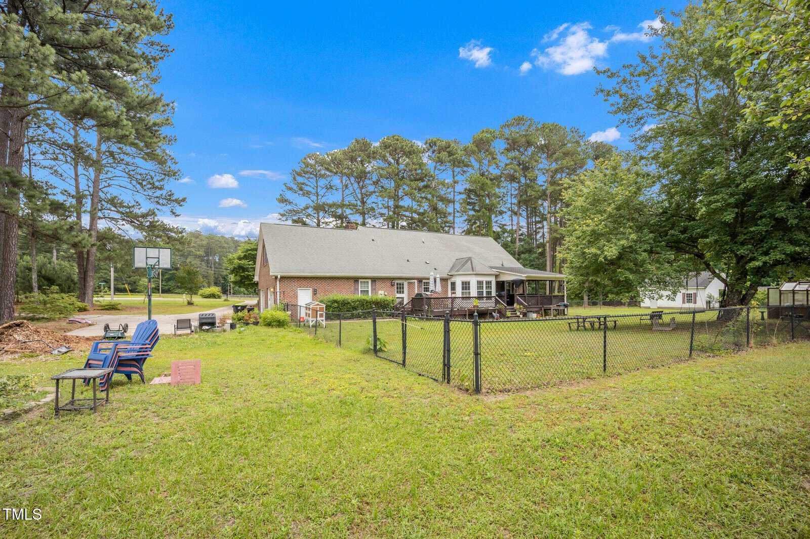 A fenced backyard with green grass, outdoor seating, and a fire pit. A brick house with a deck is in the center. Pine trees, a basketball hoop, and a clear blue sky are in the background.