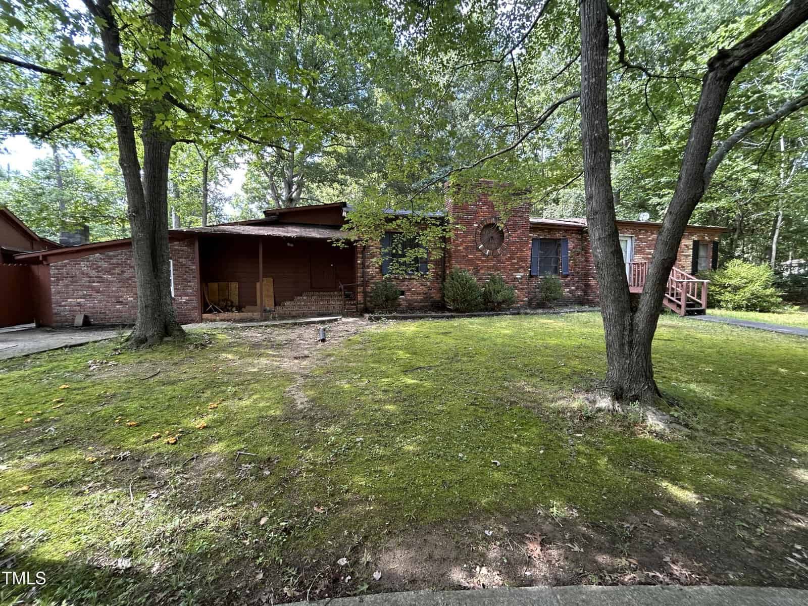 Single-story brick house with a covered porch and wooden steps, surrounded by tall trees and a grassy yard. The home is set back from the street, with scattered sunlight filtering through the leaves.
