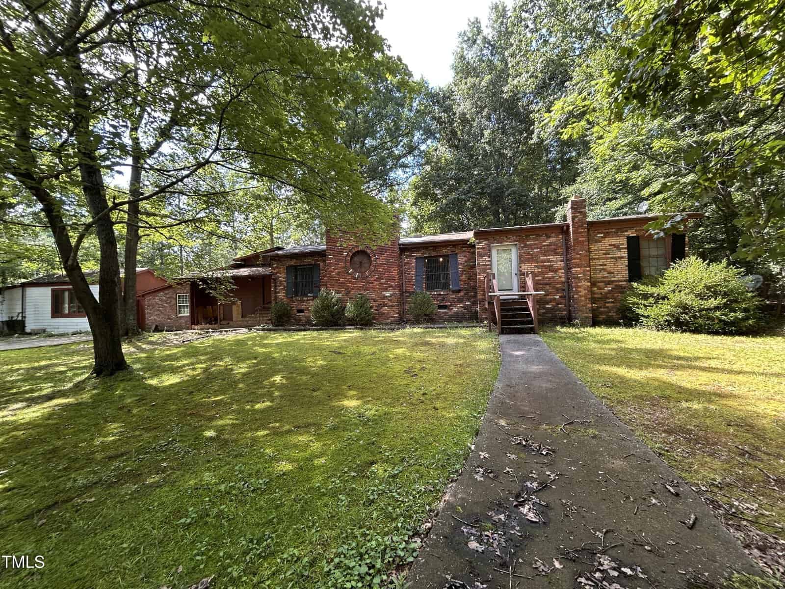 A single-story brick house with dark shutters and a central circular window, surrounded by trees and a grassy yard. A concrete pathway leads to steps at the front door. Sunlight filters through the trees.
