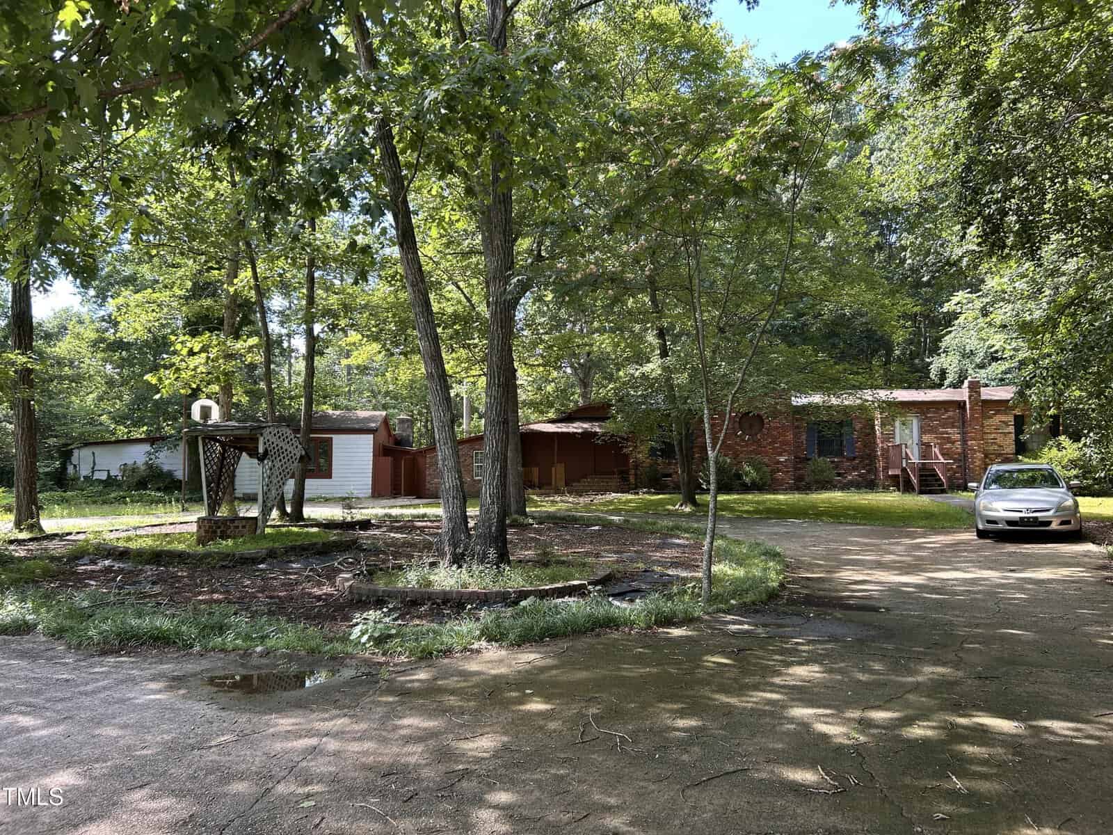 A brick house partially hidden by trees sits beside a curved driveway. A silver car is parked on the right, and a wooden structure stands on the left near grassy and wooded areas. Sunlight filters through the trees.