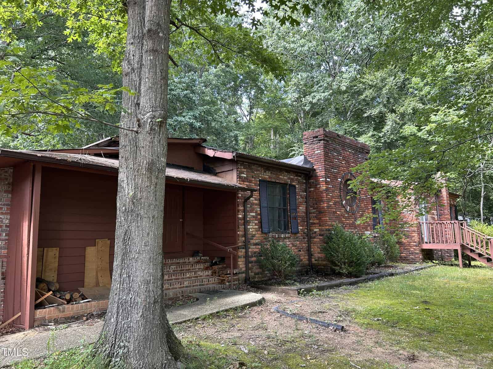 A rustic brick house with a chimney, wooden porch, and steps, surrounded by green trees and grass. Some firewood is stacked near the porch, and the area is shaded by large tree branches.