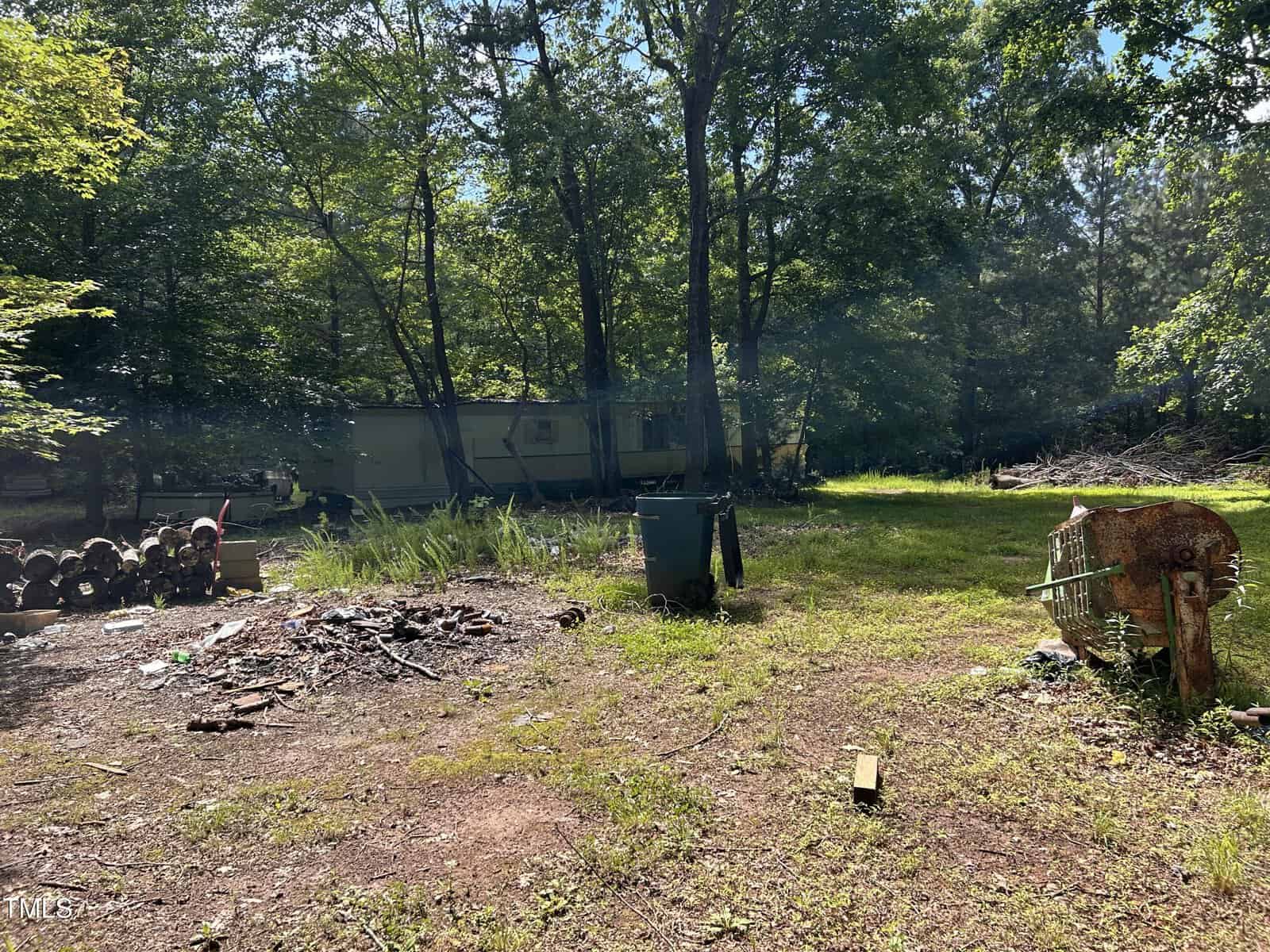 A trailer sits among tall trees in a clearing. In the foreground are a pile of firewood, a green trash bin, a rusty barrel, and scattered debris on the ground. Sunlight filters through the leaves.