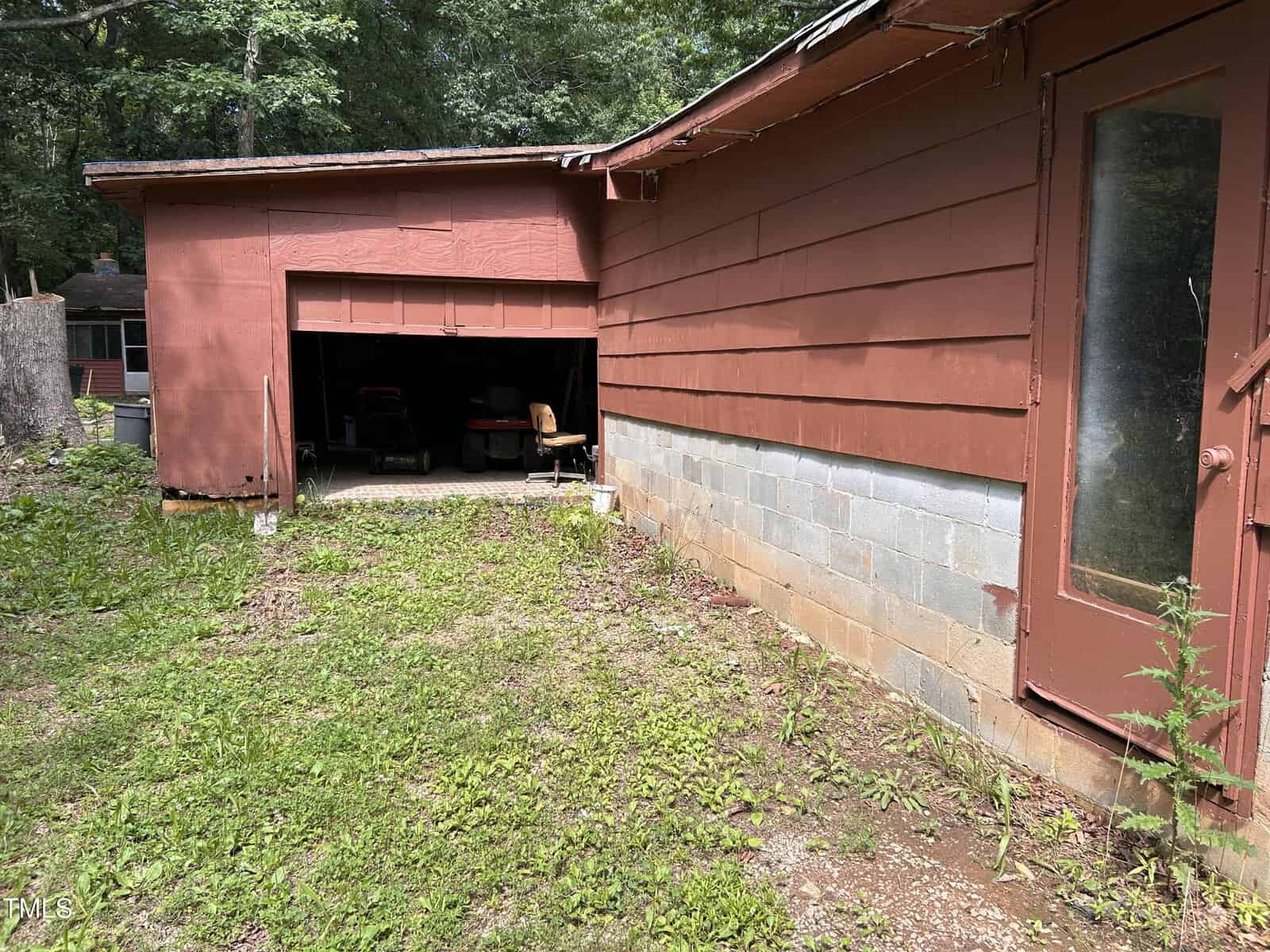 A partially open garage door reveals a lawn mower and chair inside. The exterior is red siding with a glass door and visible cement blocks at the base. Grass and patches of bare ground are in the foreground. Trees are in the background.