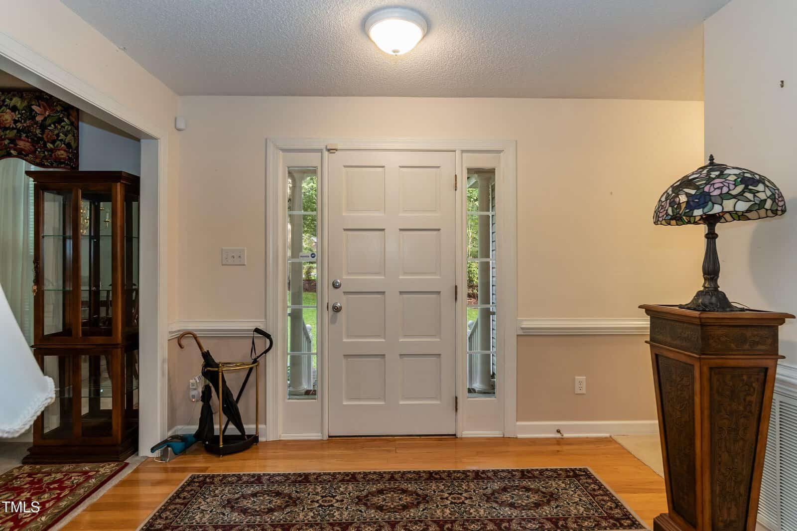 Front entryway with a white door, two tall side windows, umbrella stand, ornate cabinet, and a Tiffany-style lamp. Wood floor and patterned rug cover the space. A glass display cabinet is visible in the adjacent room.