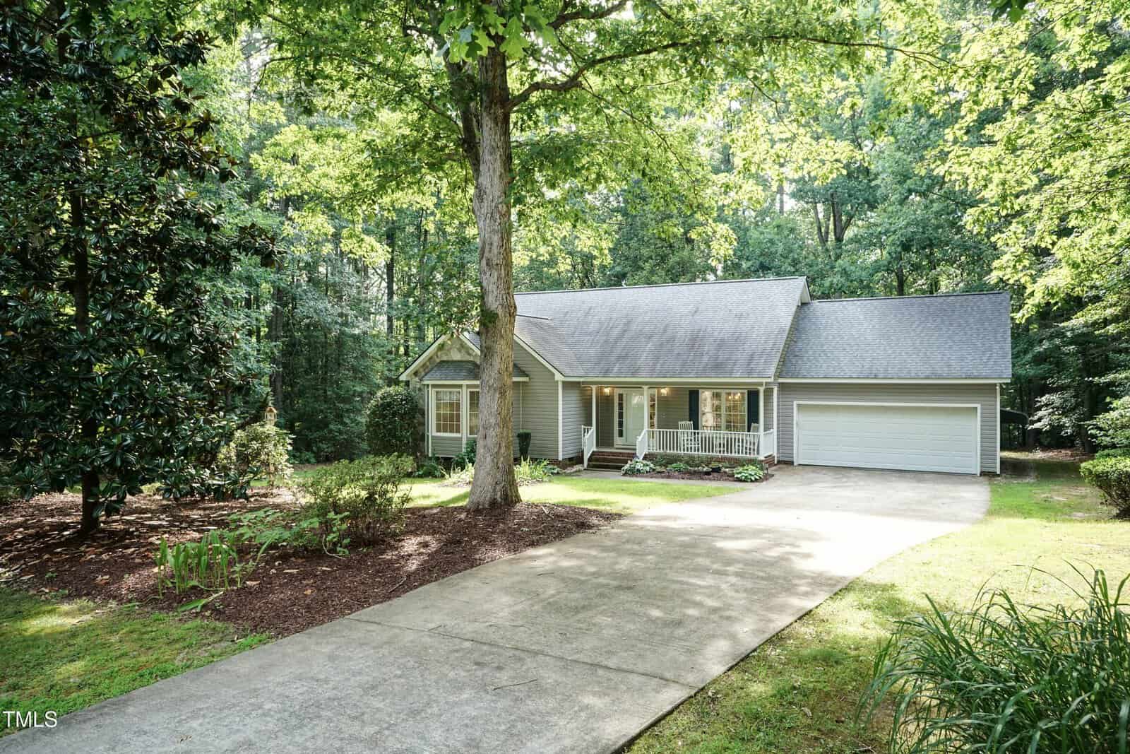 A light gray house with a front porch and attached two-car garage, set among tall trees with a large driveway and green lawn in a wooded, suburban area.