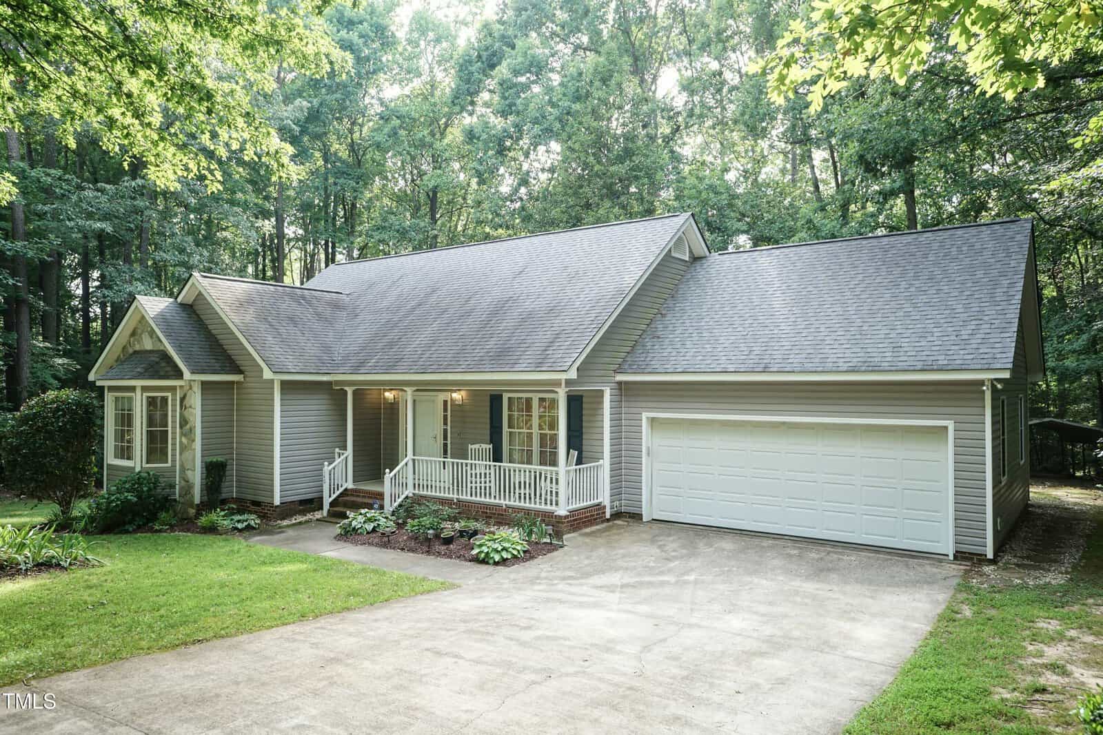 A single-story gray house with white trim, a covered front porch, and a two-car garage sits surrounded by tall trees and greenery, with a concrete driveway leading up to the house.