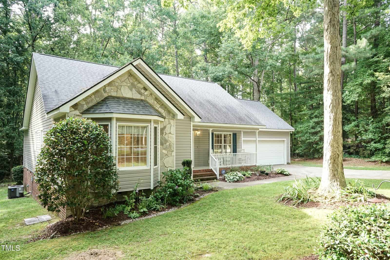 Single-story house with light-colored siding, a gabled roof, and stone accents. There is a front porch with white railing, a two-car garage, landscaped yard, and trees in the background.
