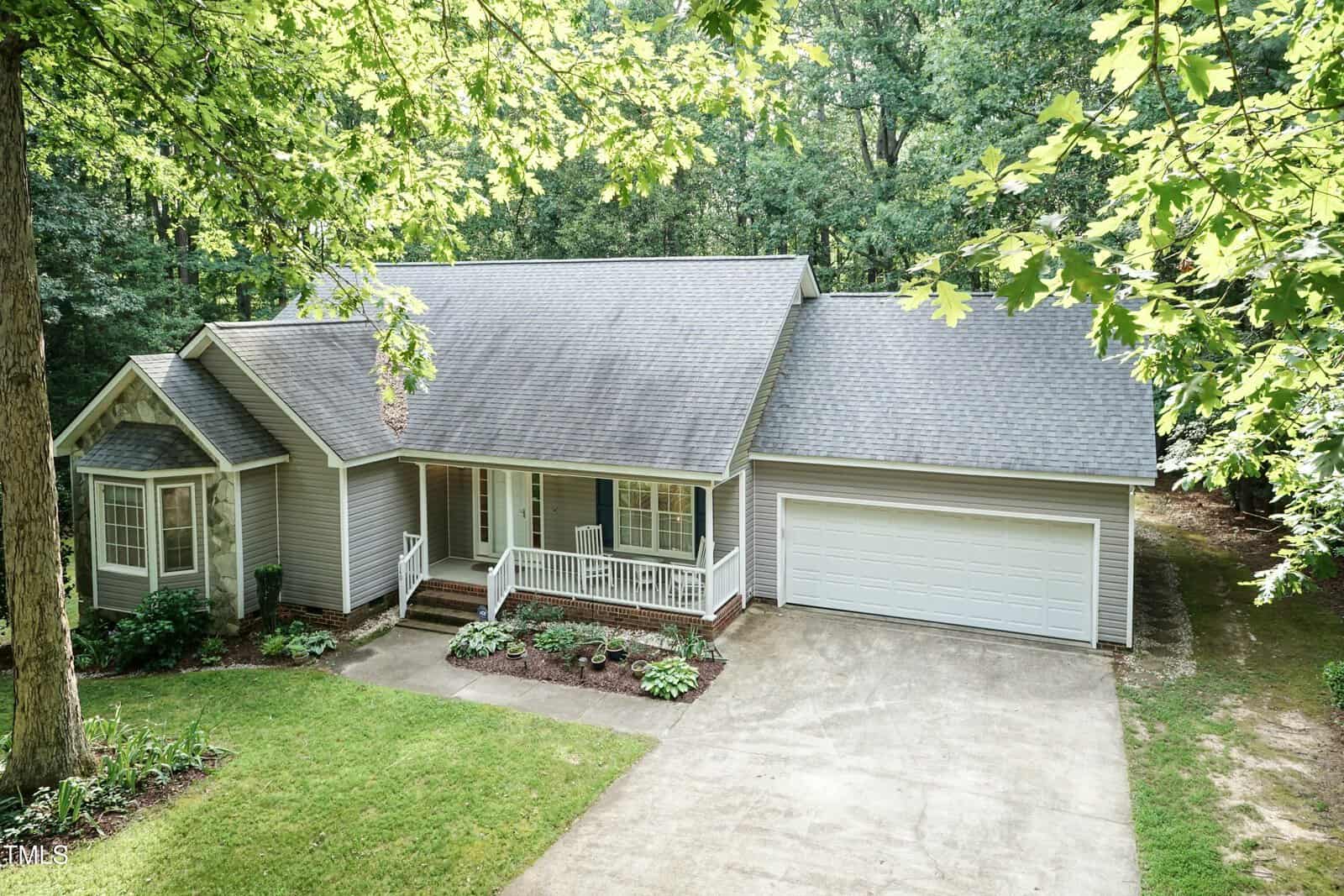 A gray, single-story house with a covered front porch, white trim, and an attached two-car garage. The home is surrounded by trees and has a neat lawn and a concrete driveway.