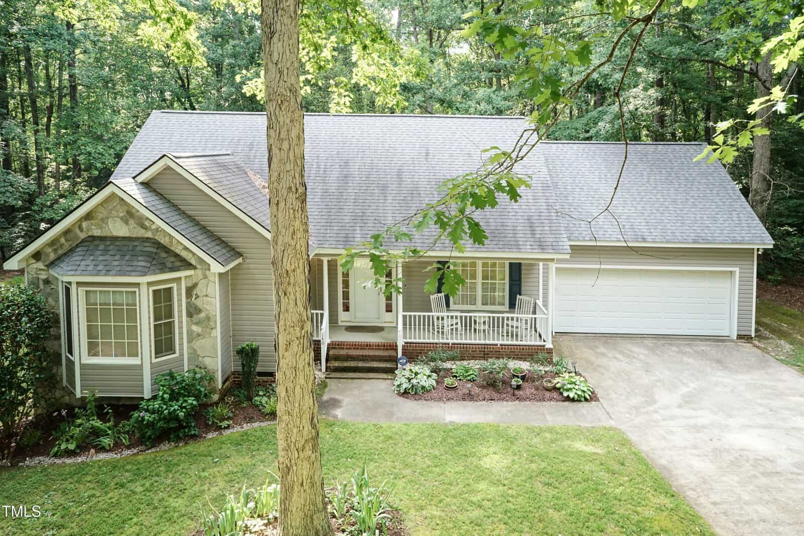 A light gray single-story house with a front porch, two-car garage, and bay window, surrounded by trees and a well-kept lawn. A large tree stands in the front yard.
