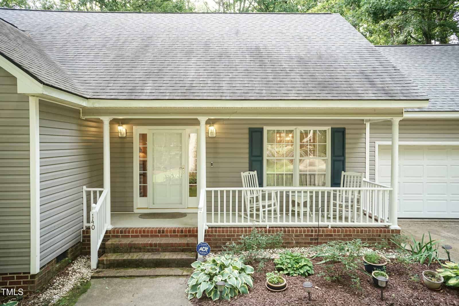 A cozy house front with a covered porch, two white rocking chairs, potted plants, and a garden bed. The house has light gray siding, a white door with glass panes, and a one-car garage.
