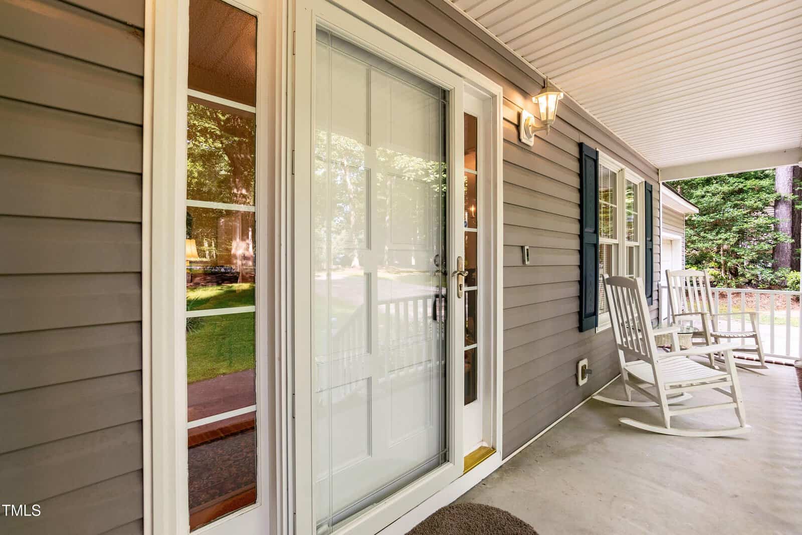 A covered front porch with two white rocking chairs, a glass storm door, beige siding, a wall-mounted exterior light, and a view of trees and lawn reflected in the glass. White railings and trim are visible.