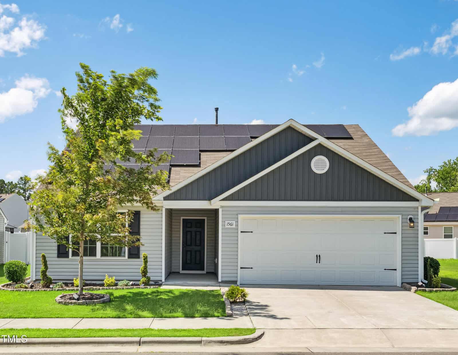 A single-story house with gray siding, a gabled roof with solar panels, a double garage, and a black front door. A small tree and neatly trimmed grass are in the front yard under a blue sky with light clouds.