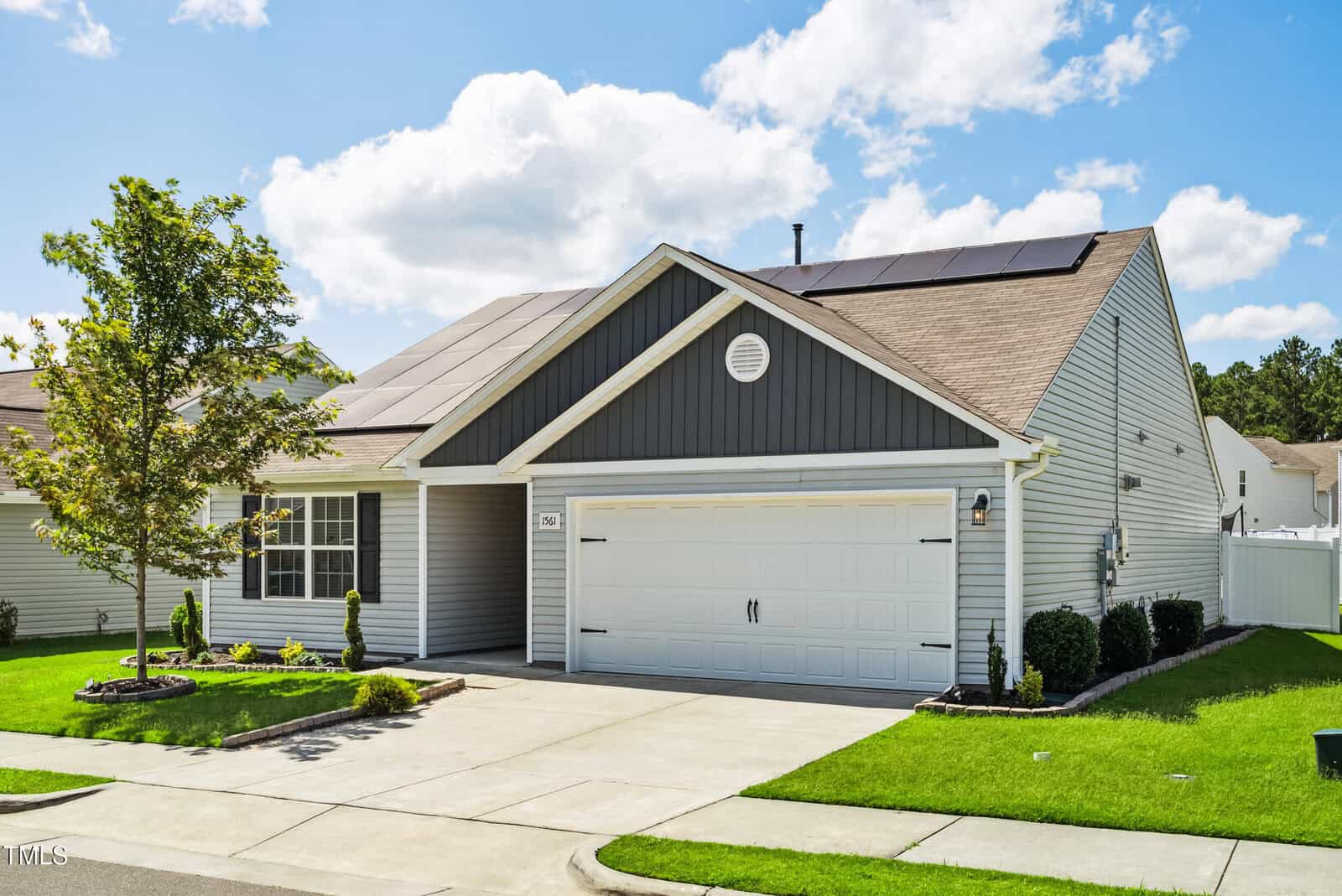 A single-story house with gray siding, white trim, a double garage, and solar panels on the roof. The front yard has green grass, small shrubs, a tree, and a concrete driveway under a partly cloudy sky.