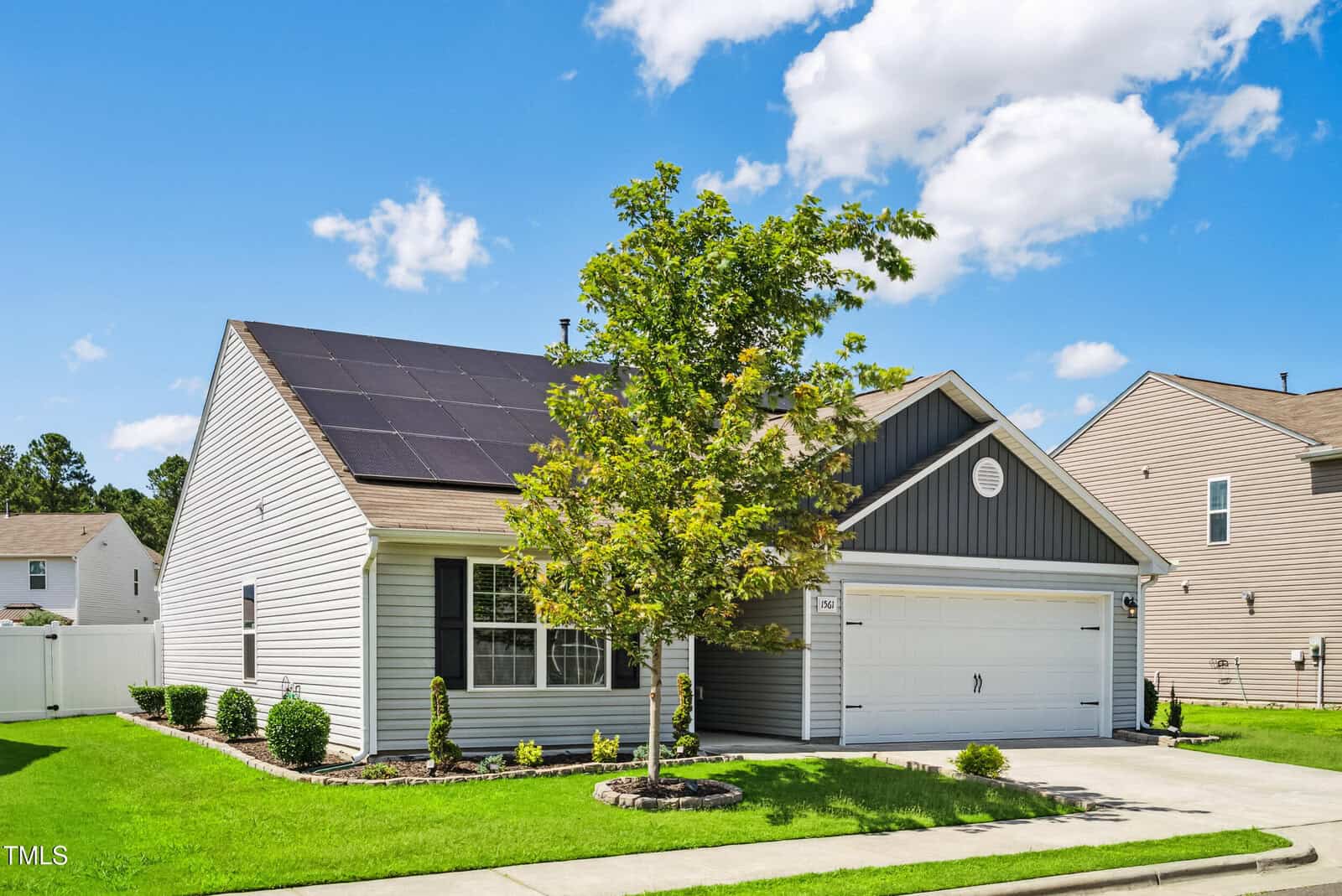 Single-story house with gray siding, white trim, and solar panels on the roof. A tree partially obscures the front, with a driveway leading to a two-car garage. The sky is blue with scattered clouds.