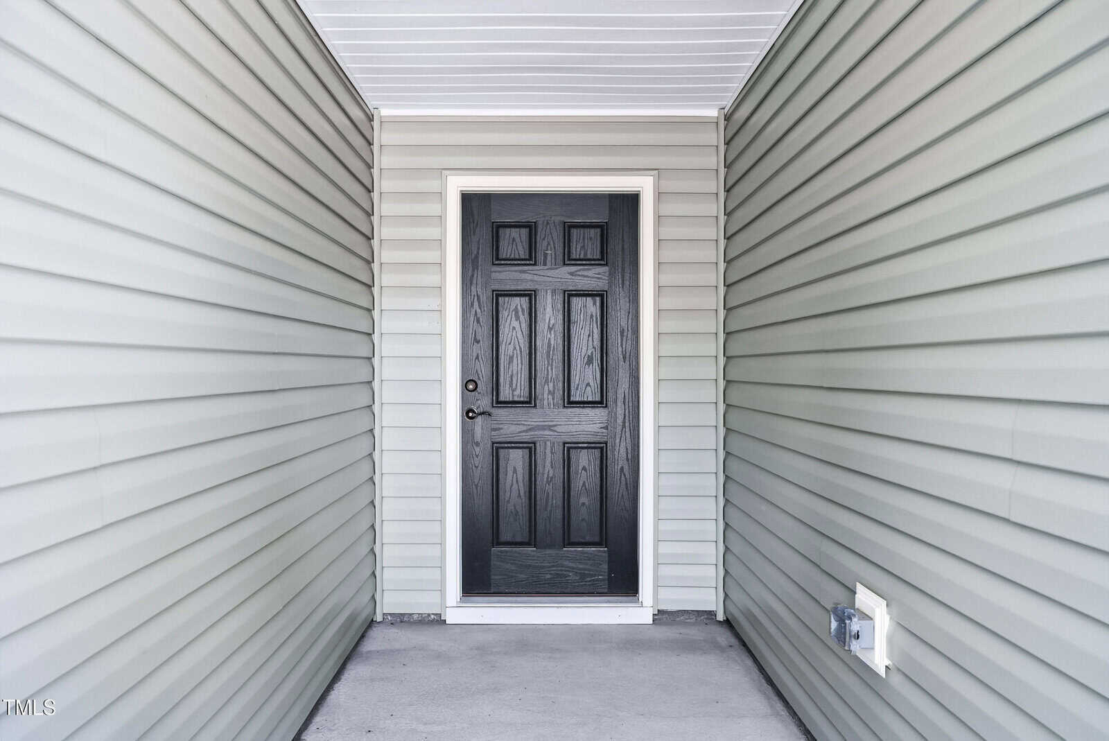 A black front door with six panels is centered between light gray vinyl siding walls and a concrete floor, under a white tongue-and-groove ceiling.