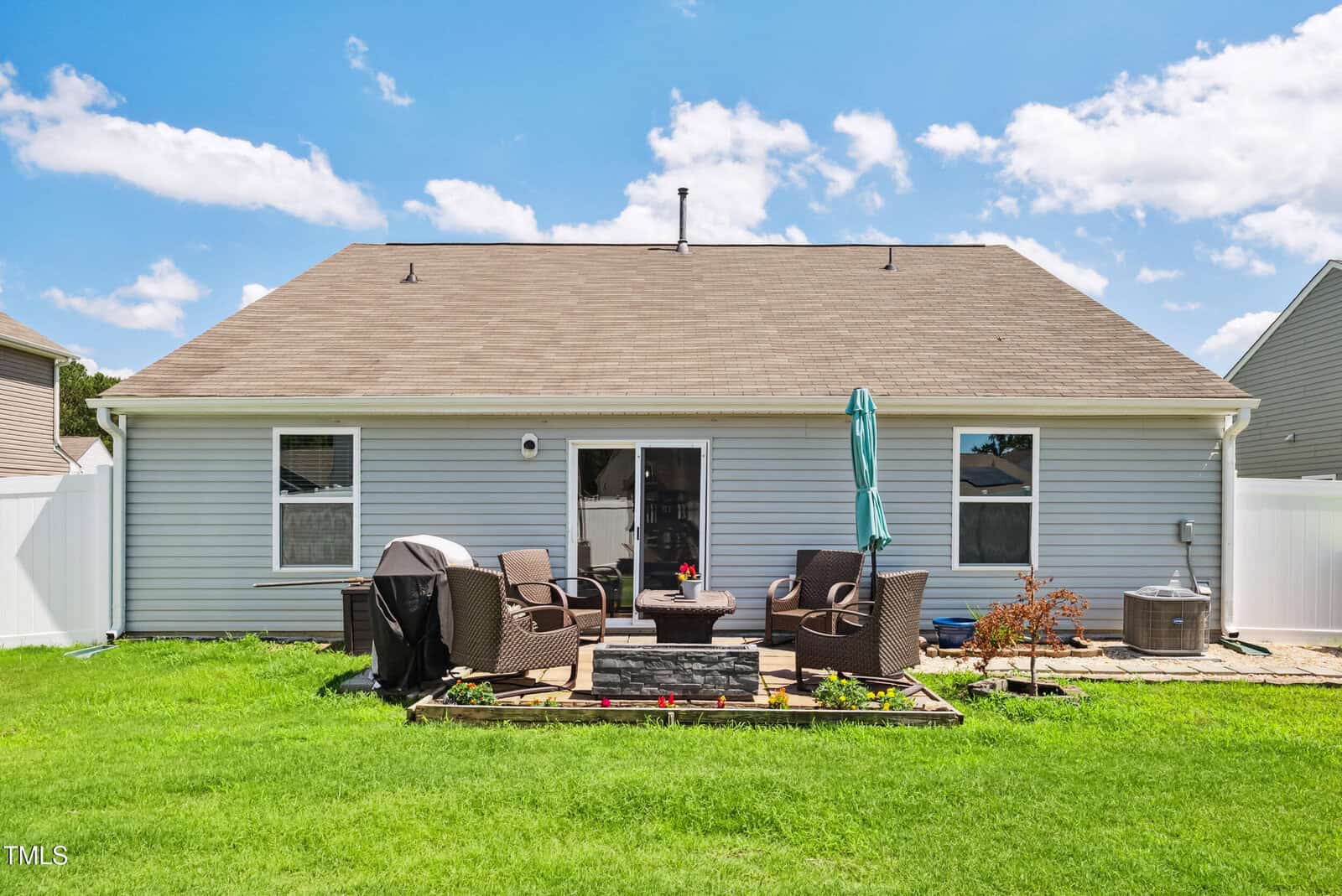 A backyard view of a single-story house with light gray siding, a shingled roof, two windows, and a glass door. The patio has wicker furniture, a grill, an umbrella, and a small fire pit, set on green grass.