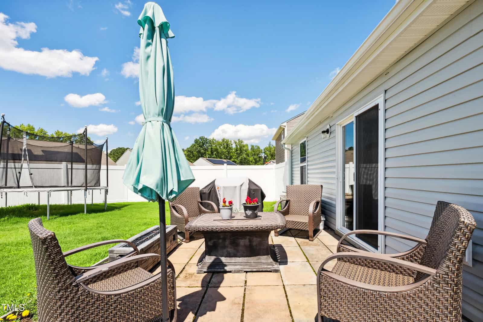 A backyard patio with four wicker chairs around a square table, a closed teal patio umbrella, and potted plants on the table. The patio is next to a house with gray siding and sliding glass doors. Grass lawn and trampoline in background.