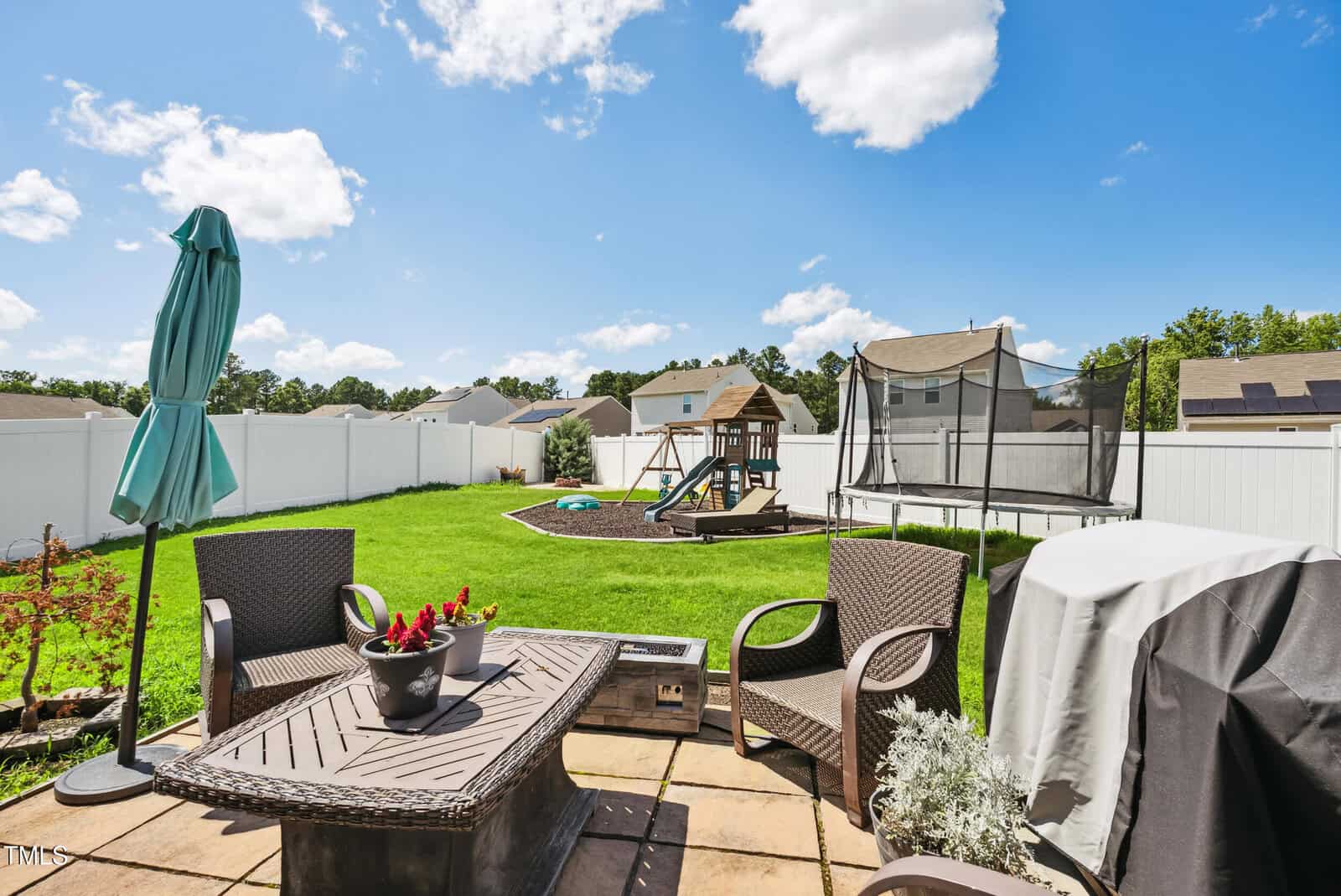 A backyard patio with four wicker chairs and a table with potted plants, a covered grill, and an umbrella. The yard has green grass, a playset, a trampoline, and is enclosed by a white fence.