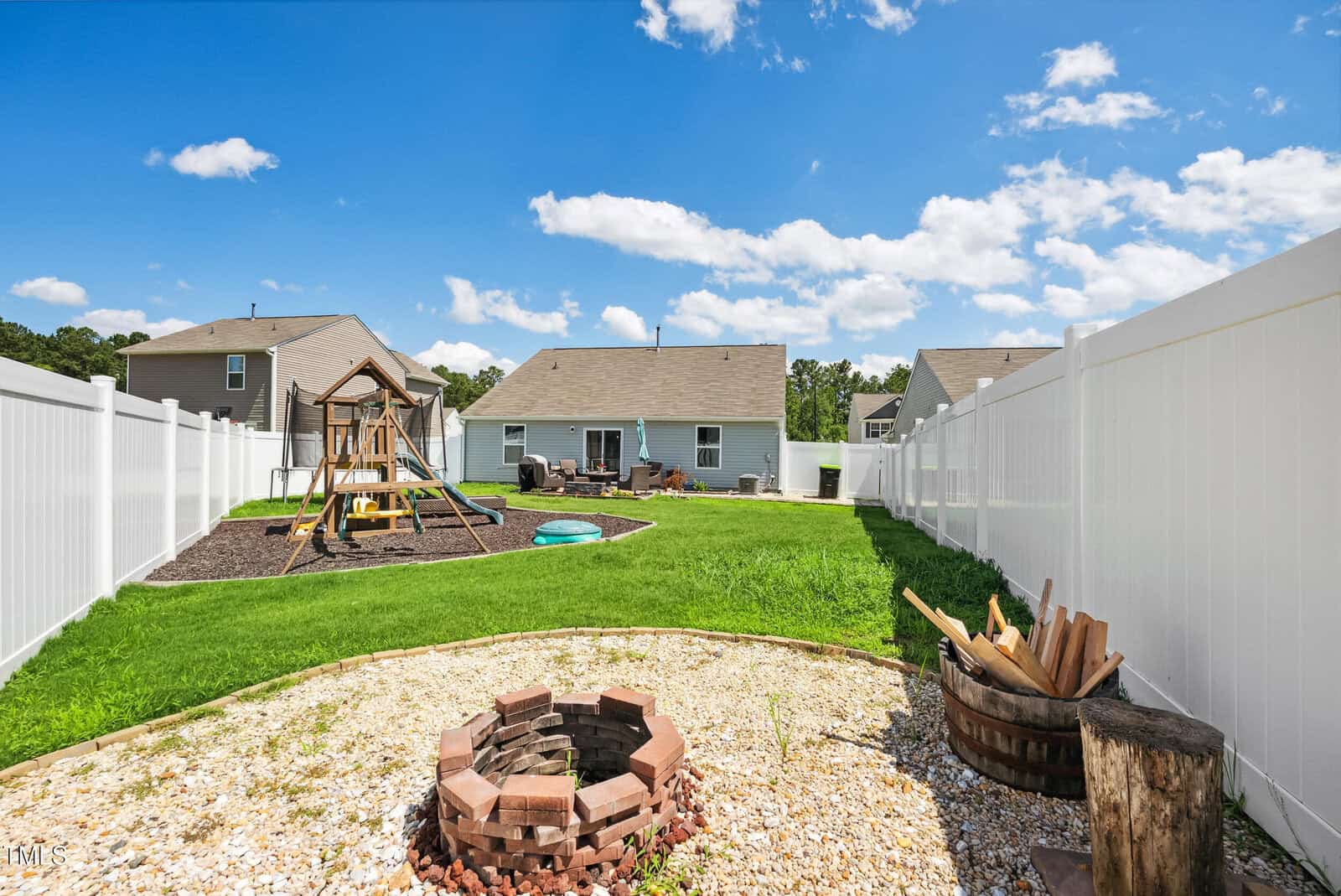 A backyard with white vinyl fencing, a playset with slides, a patch of green grass, a circular fire pit lined with bricks, gravel surrounding the fire pit, and a wooden barrel filled with firewood under a blue sky.
