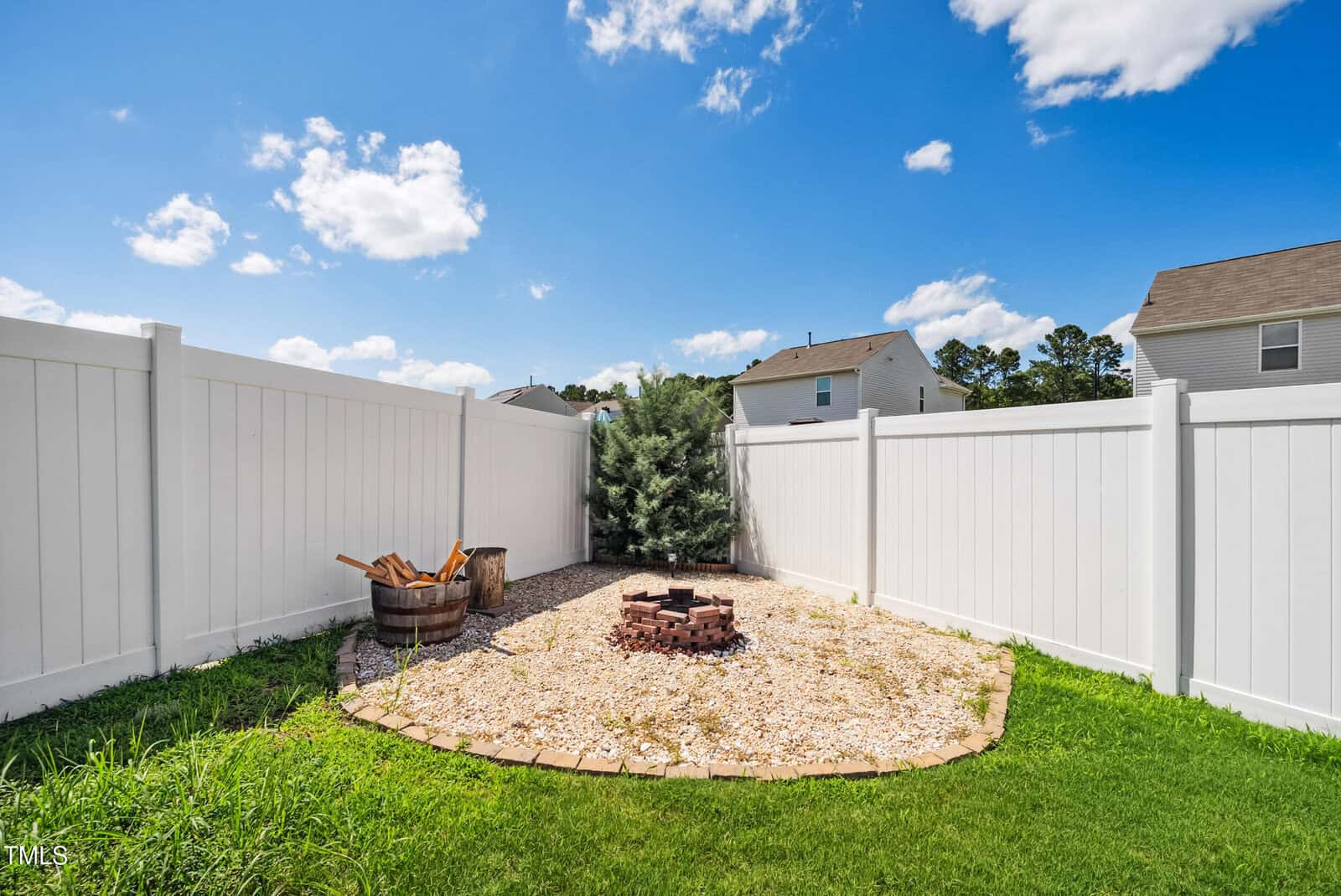 A small backyard area enclosed by a white vinyl privacy fence, featuring a circular gravel section with a brick fire pit, a wooden barrel with firewood, and a green bush in the corner under a blue sky.