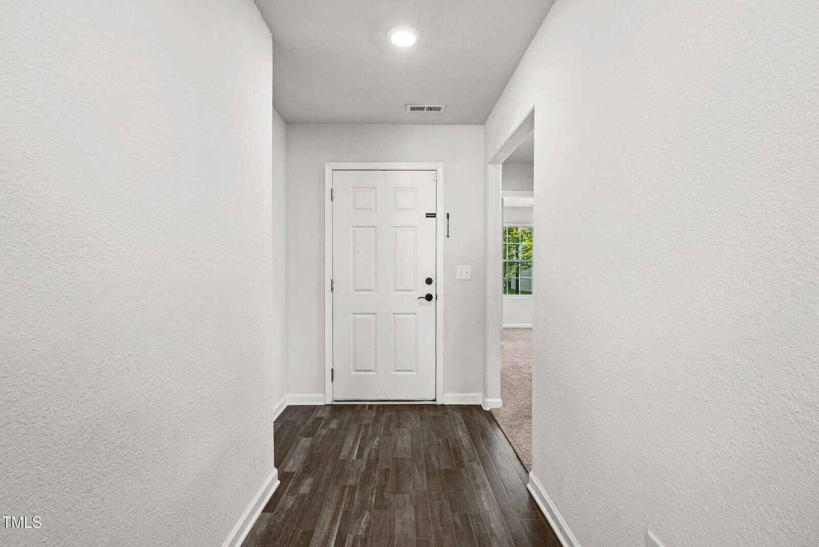 A hallway with dark wood flooring leads to a white front door. The walls are white, and a doorway on the right opens to a carpeted room with a window. A ceiling light is above the entryway.
