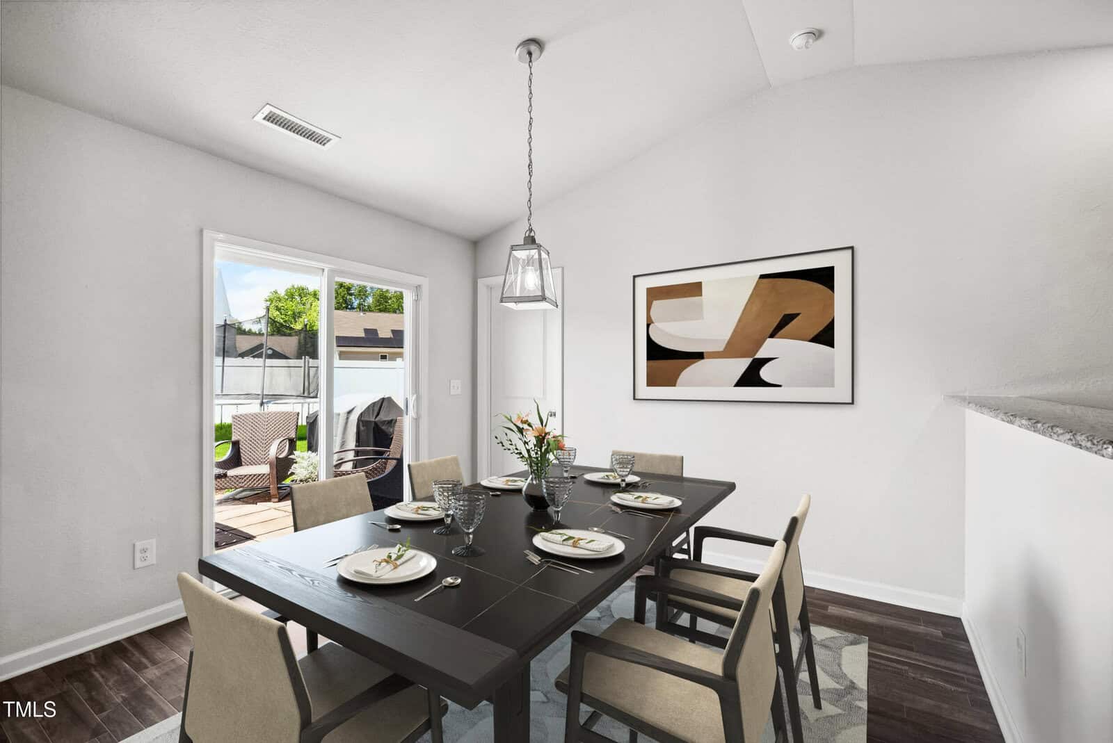 Modern dining room with a black table set for six, beige chairs, abstract art on the wall, and sliding glass door opening to a patio with outdoor furniture. White walls and dark wood flooring.