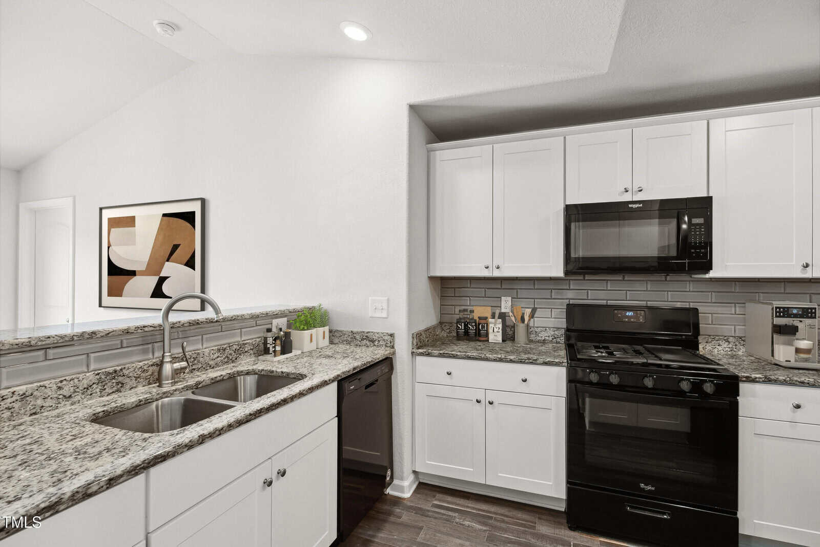 Modern kitchen with white cabinets, granite countertops, a double sink, black gas stove, black microwave, gray subway tile backsplash, and abstract wall art. Dark wood floor contrasts with the bright, clean decor.