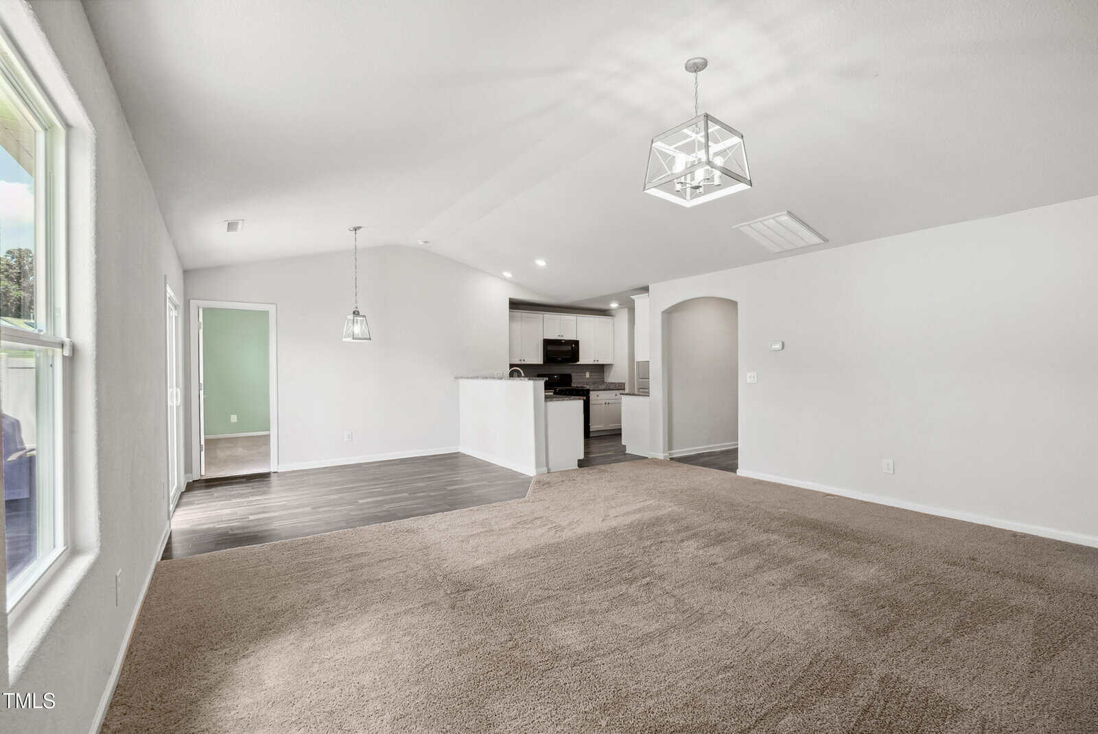A bright, unfurnished open-concept living area with beige carpet, white walls, and a view into the kitchen and dining space. The kitchen has dark flooring, white cabinets, and modern light fixtures.