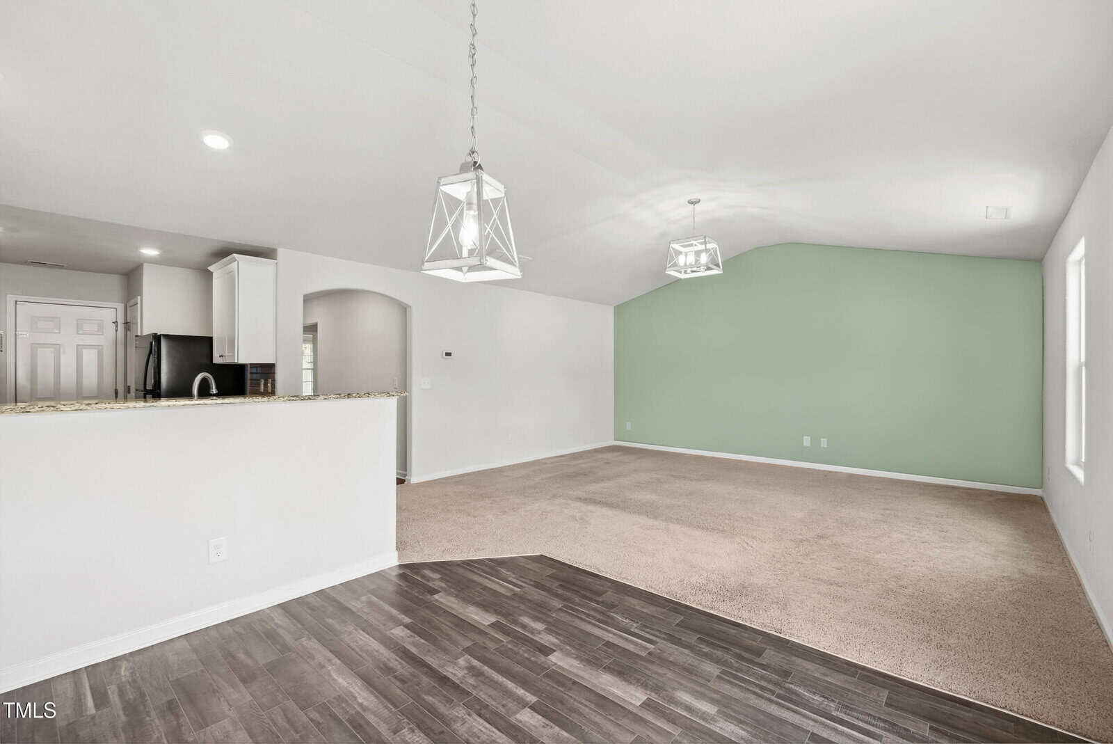 Open-concept living space with dark wood flooring leading to carpeted area, green accent wall, two geometric pendant lights, and a kitchen with white cabinets and granite countertops visible through an arched opening.