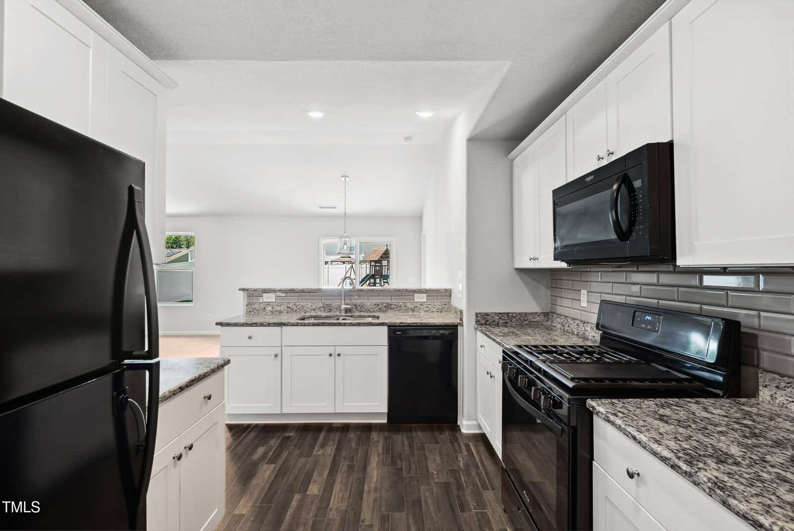 Modern kitchen with white cabinets, black appliances, granite countertops, gray subway tile backsplash, and dark wood floors. The kitchen opens to a bright dining and living area with large windows.