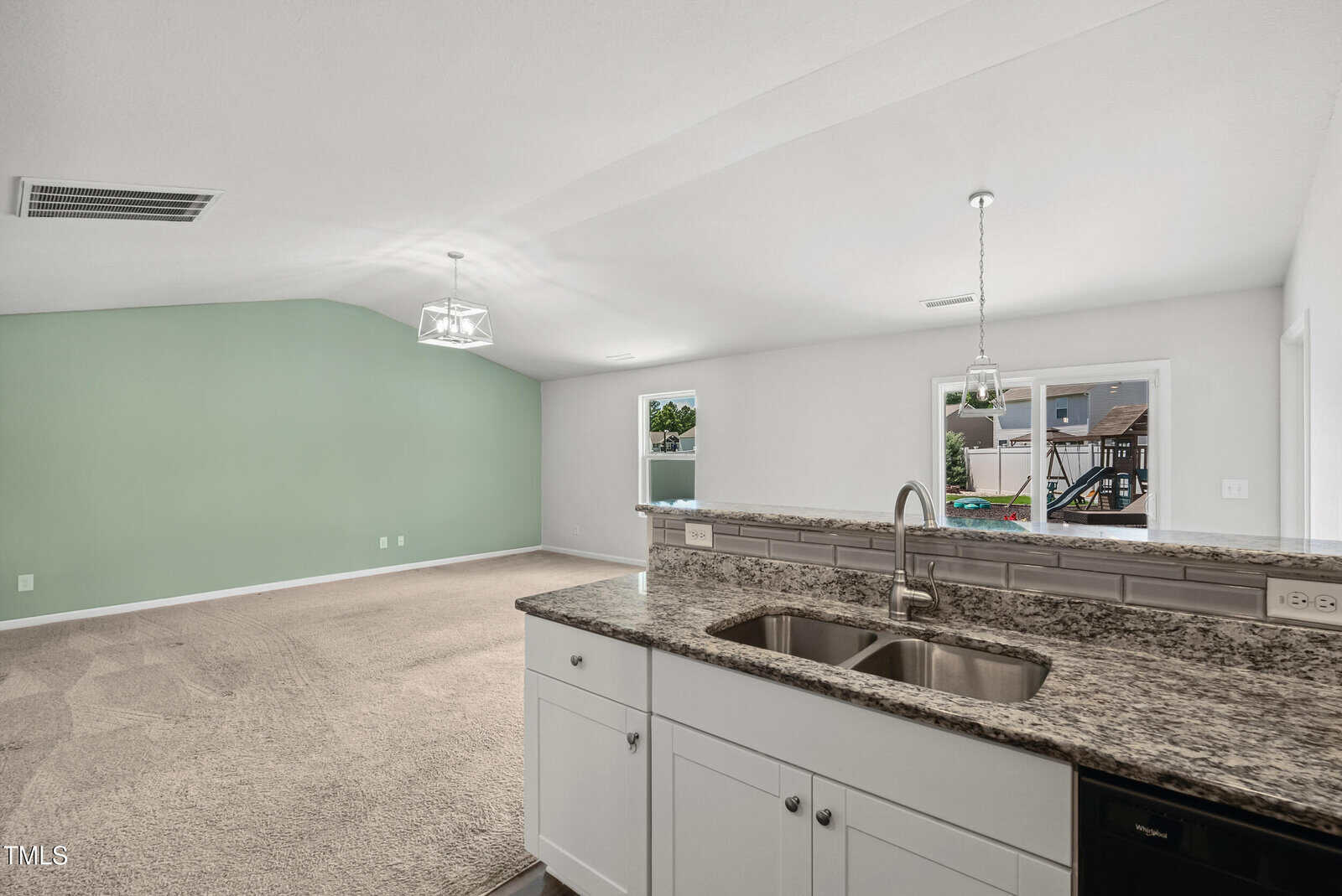 A view from the kitchen showing a double sink with granite countertops, white cabinets, and an open living area with beige carpet, a green accent wall, and windows letting in natural light.