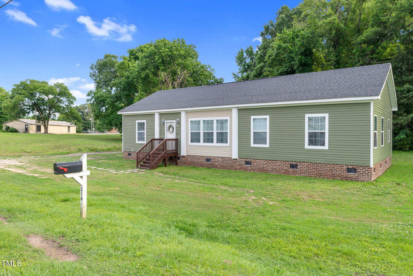 Single-story green house with white trim and a brick foundation on a large grassy lot, featuring a small porch with steps, surrounded by trees, under a blue sky with scattered clouds. A white mailbox stands in front.