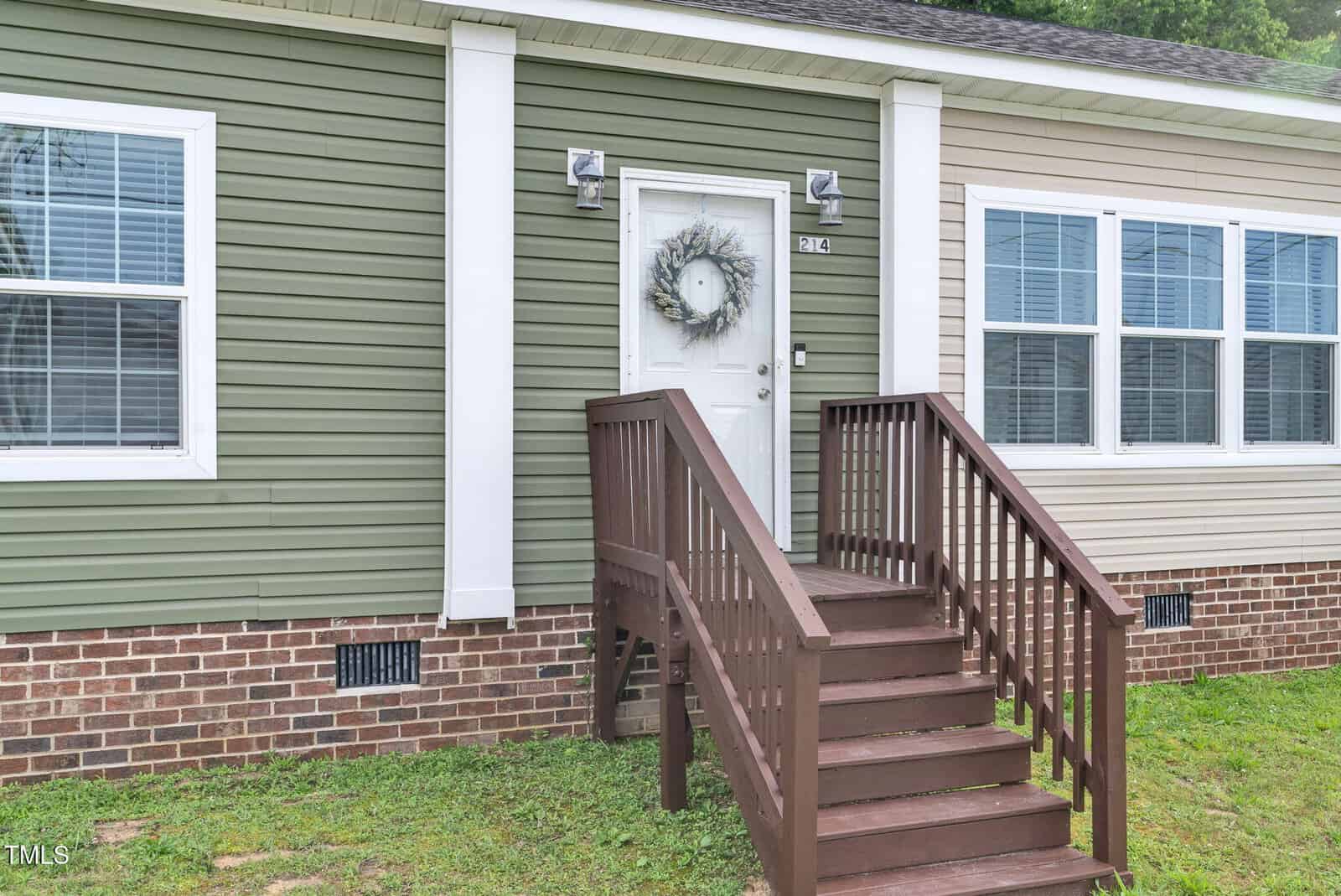 A front view of a house with green and beige siding, a white door decorated with a wreath, brown wooden steps with railings, and several windows above a brick foundation.