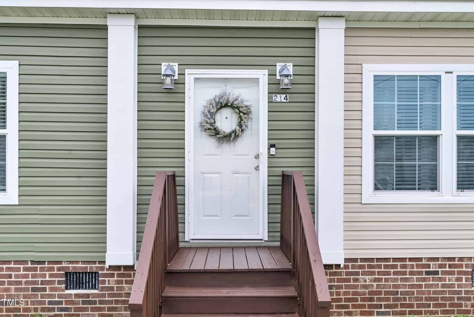 A white front door with a wreath, flanked by two wall lights, is at the top of wooden steps. The house exterior has green and beige siding with white trim and brick at the bottom. The house number 214 is visible.