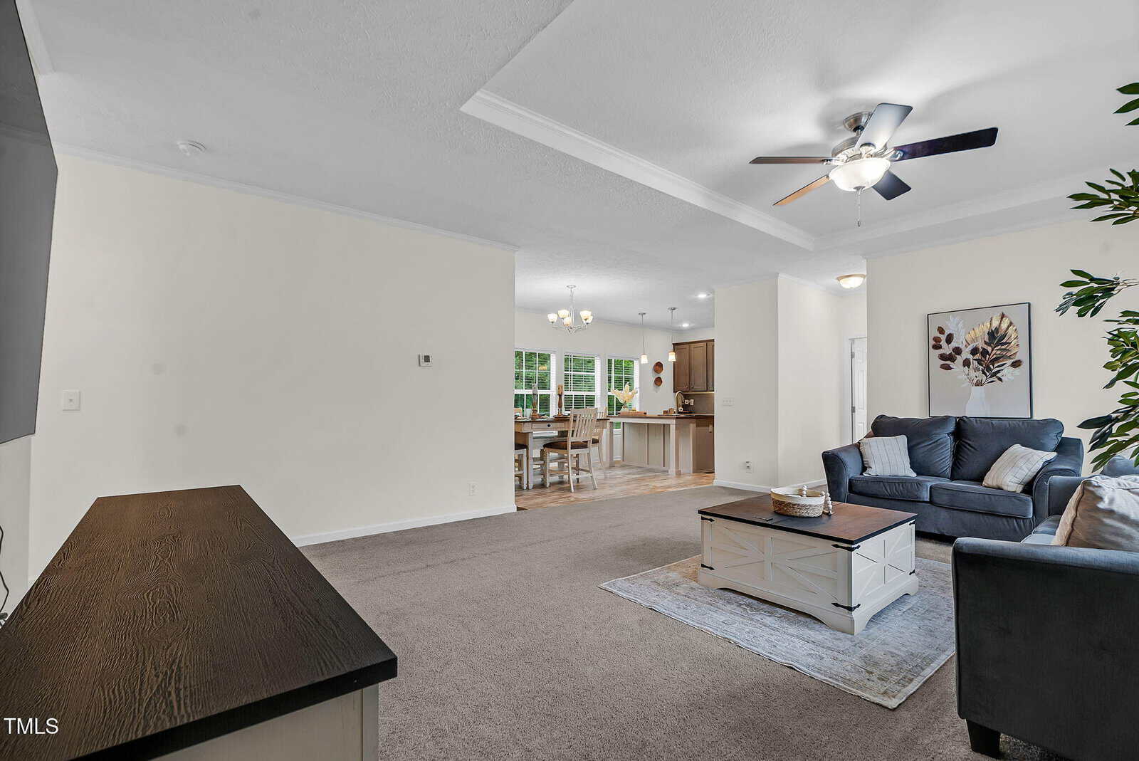 Living room with gray sofa, matching armchair, coffee table, area rug, ceiling fan, and wall art. Open layout shows dining area and kitchen with wood cabinets and a table with chairs in the background.