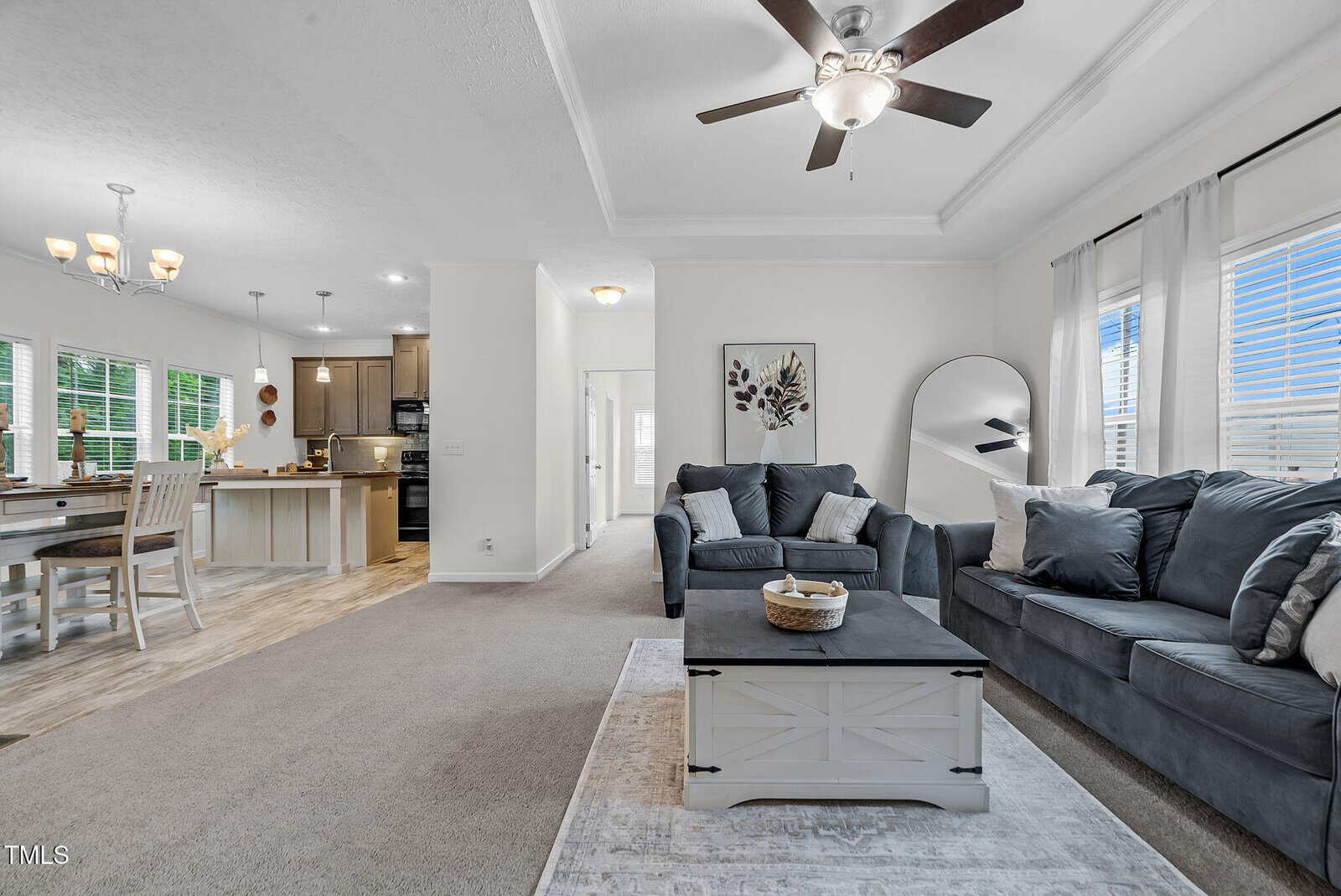 Open living area with gray sofas, a white coffee table, and a large mirror. The kitchen and dining area have light wood floors, white cabinetry, and stainless steel appliances. Ceiling fans and windows provide natural light.